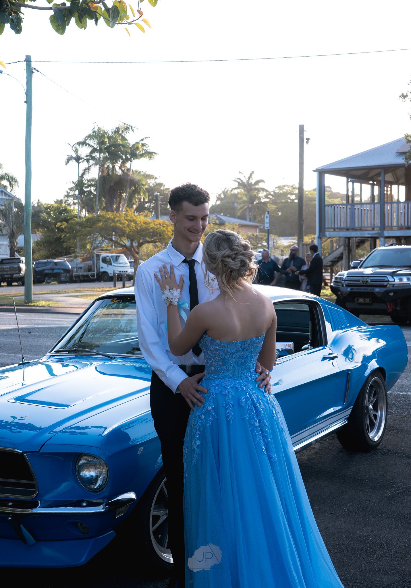 A couple in formal attire poses with a vintage blue convertible car during what appears to be a prom or special event.
