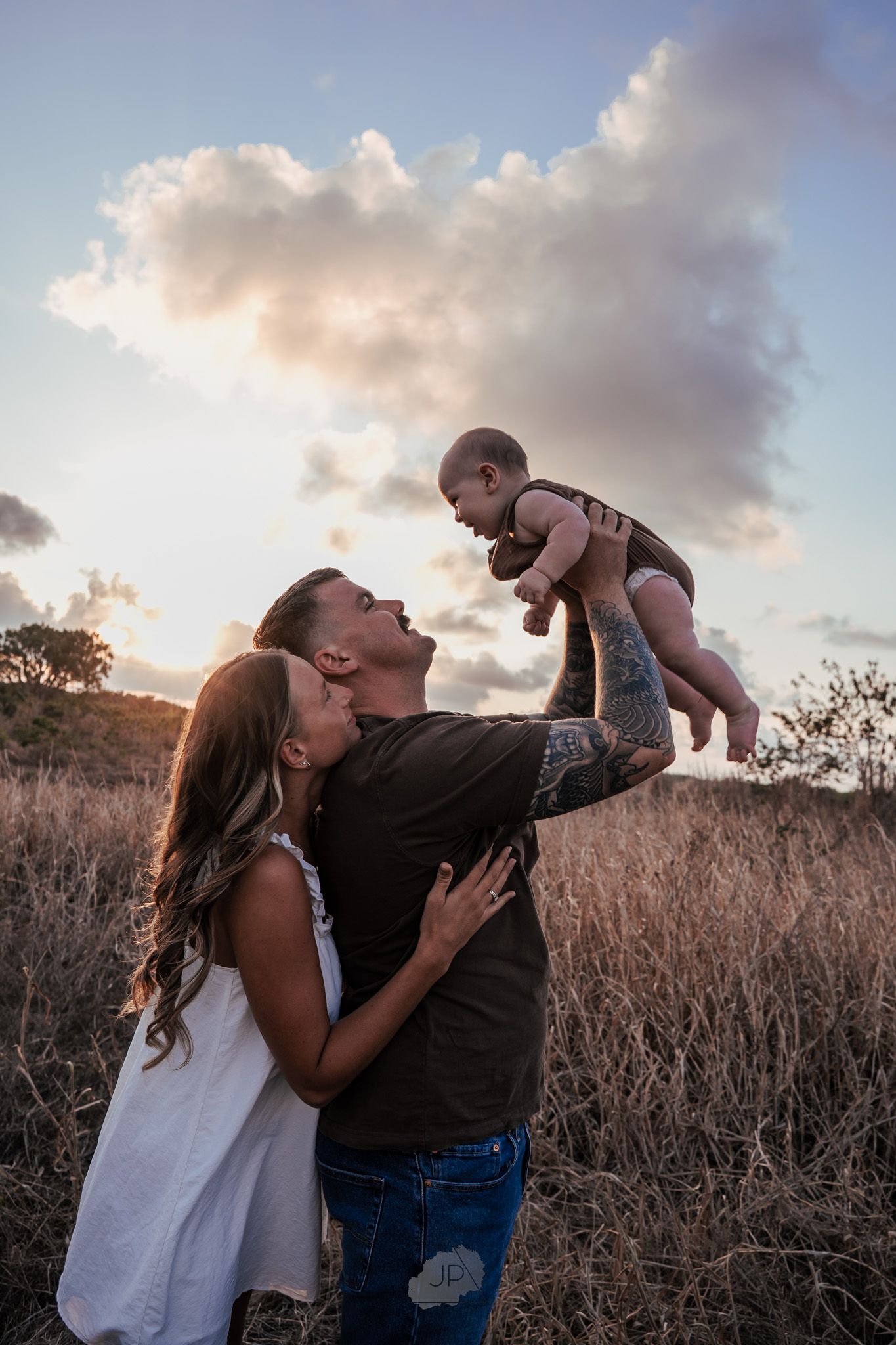 A couple shares a joyful moment with their baby in a field at sunset with dramatic clouds in the background.