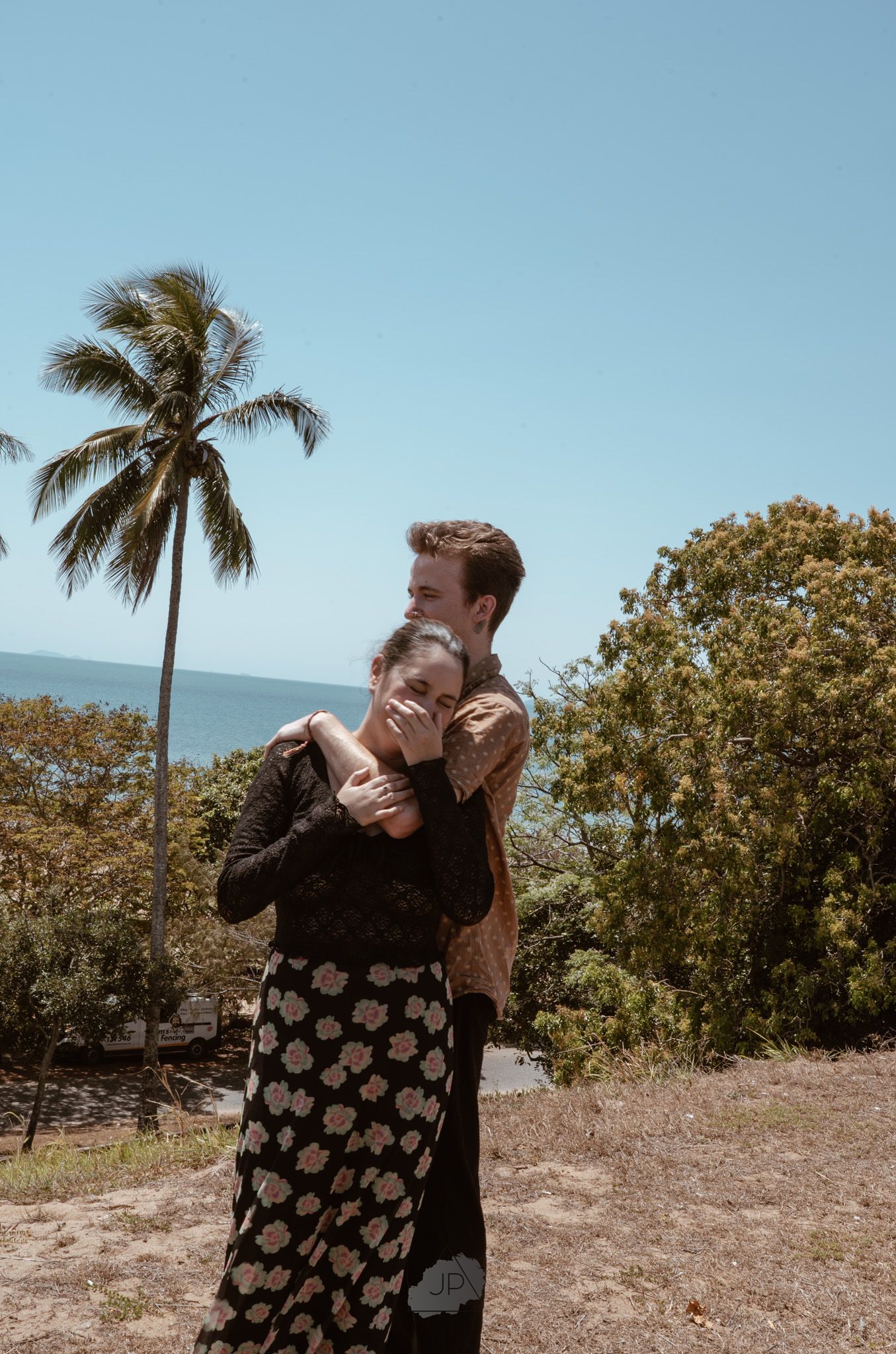 A couple embraces on a tropical beach with a palm tree and ocean view in the background.