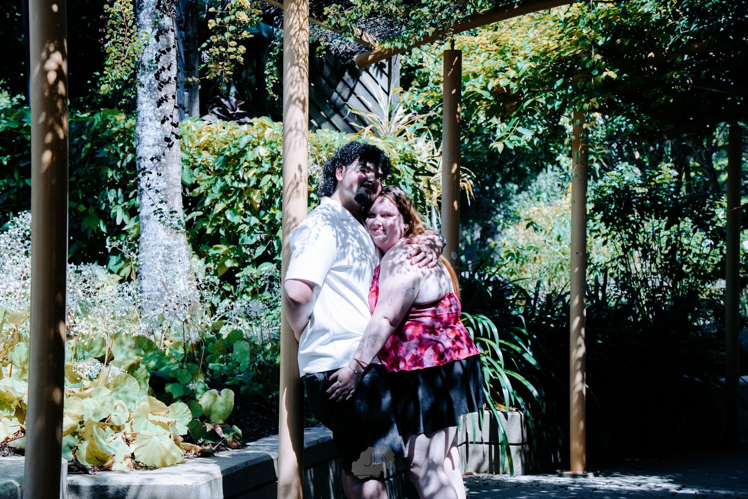 A couple stands together in a garden setting with wooden posts and lush greenery in the background.