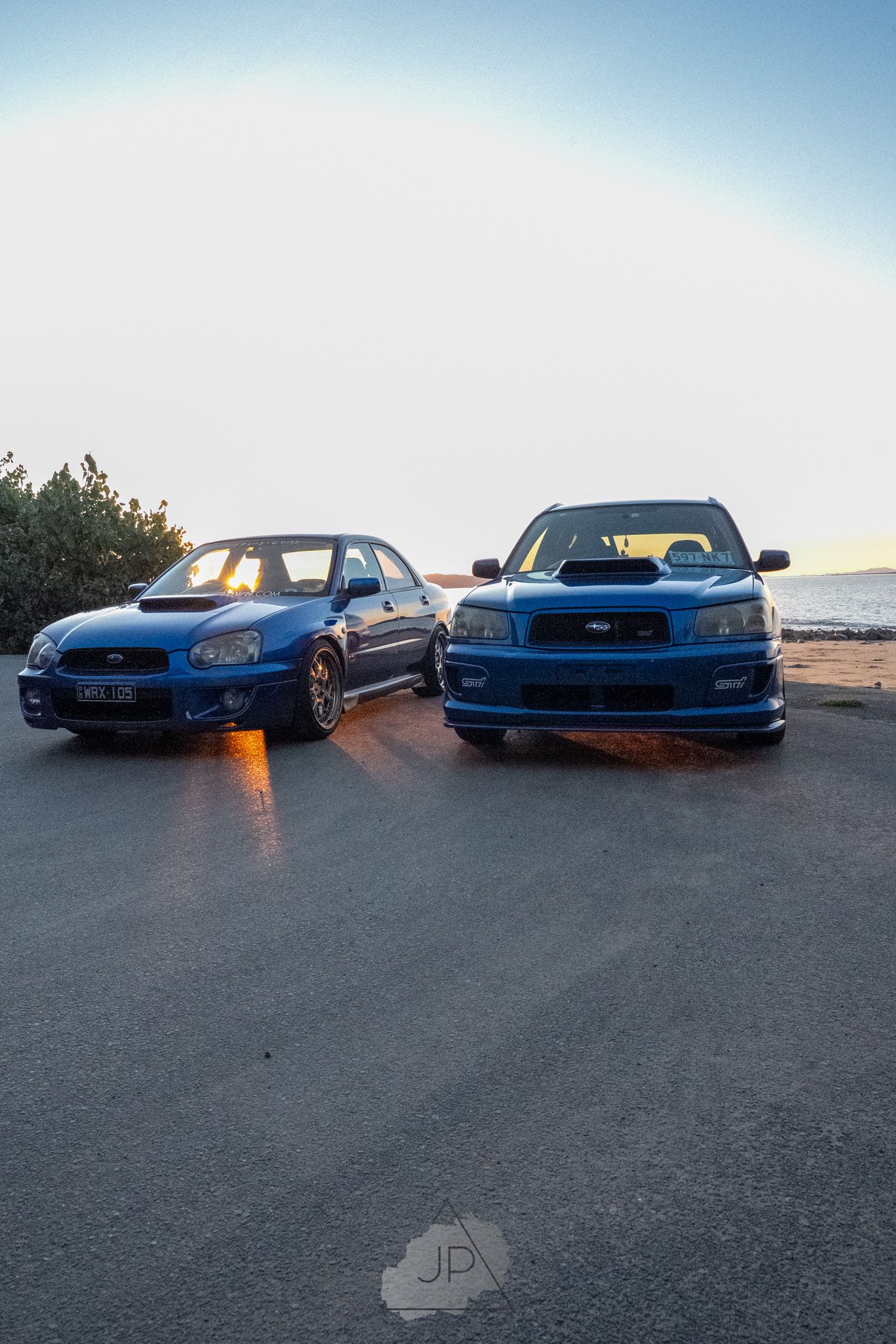 Two blue Subaru WRX STI sports cars parked side by side at sunset with parking lights illuminated.