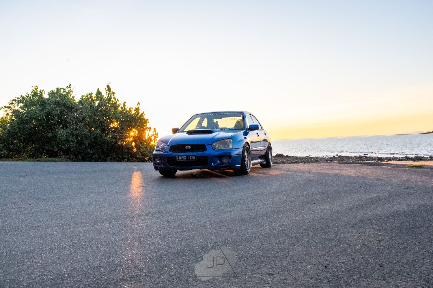 Blue compact car parked near the ocean during sunset with dramatic lighting and coastal scenery.