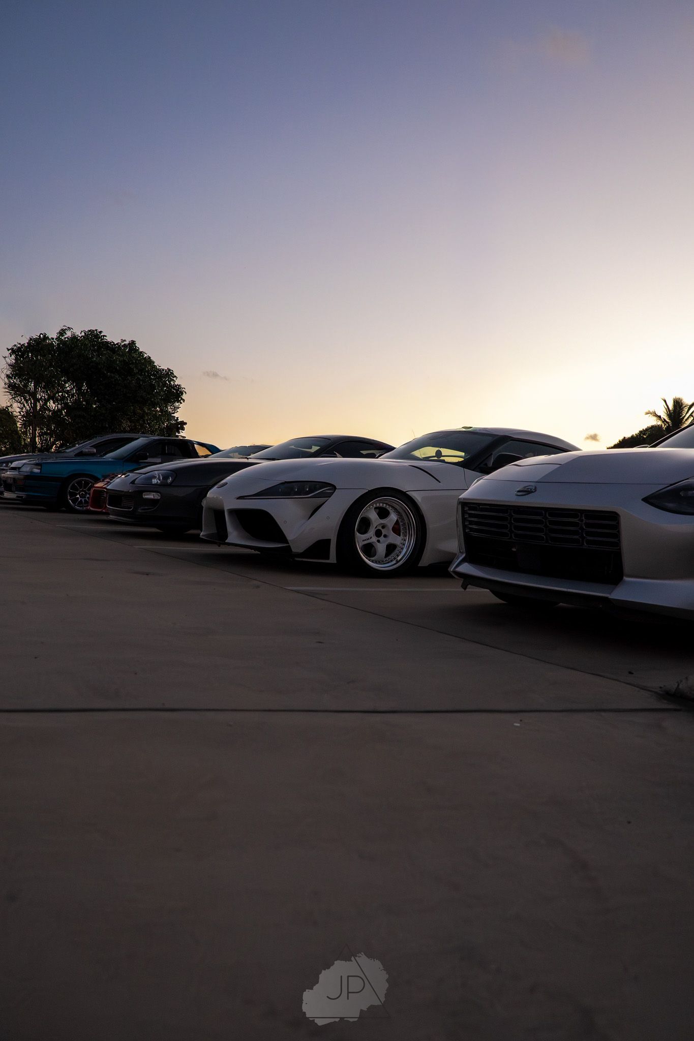 Multiple sports cars including a Toyota Supra lined up in a parking lot during golden hour sunset.