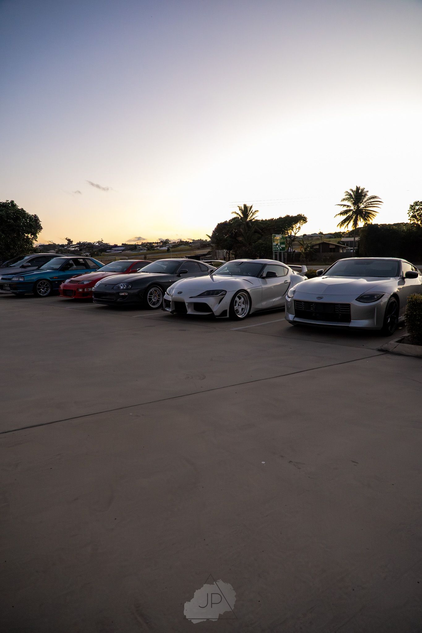 A line of luxury sports cars parked together at dusk with palm trees silhouetted against the sunset sky.