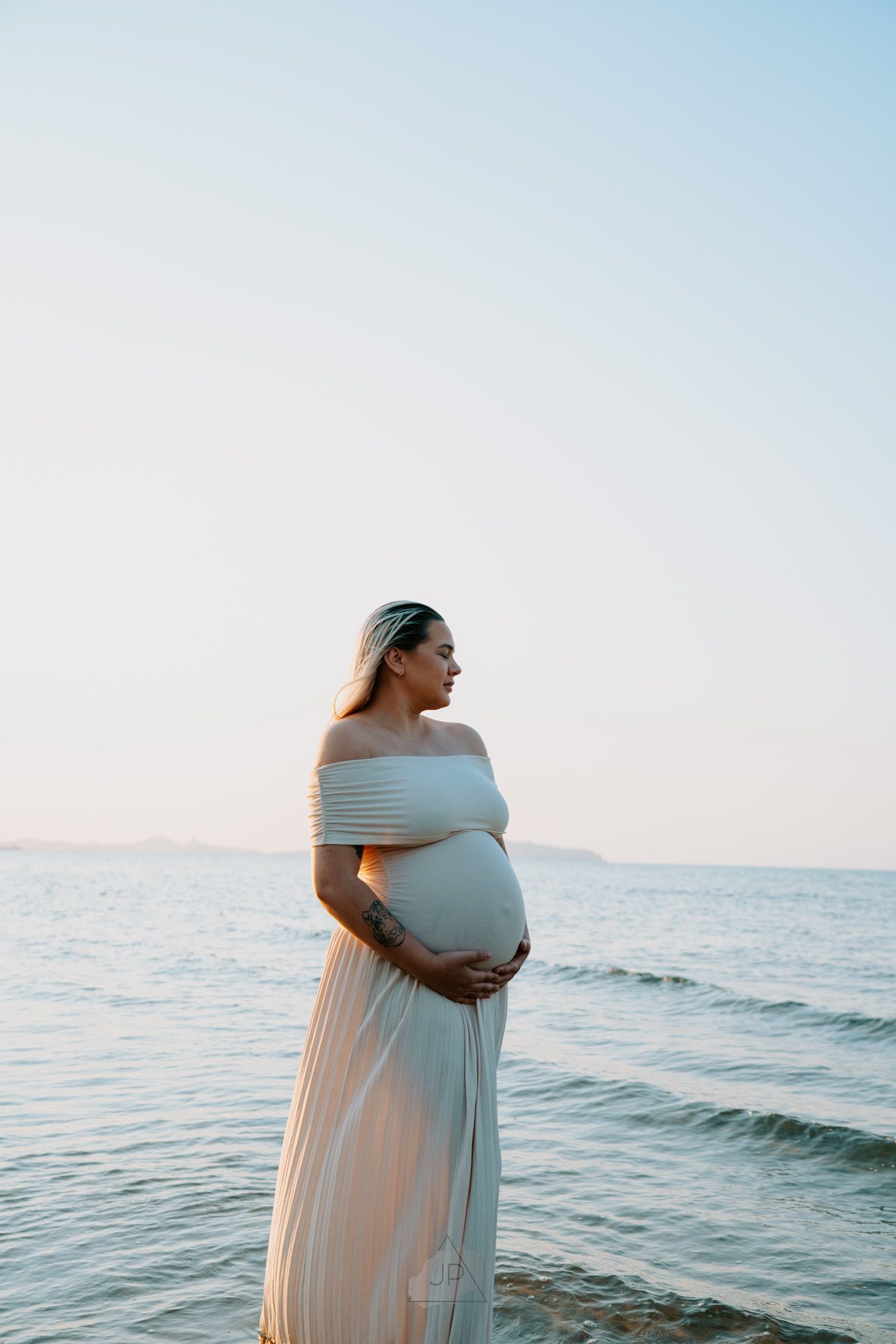 A pregnant woman in a flowing white dress stands by the ocean during sunset, creating a serene maternity portrait.
