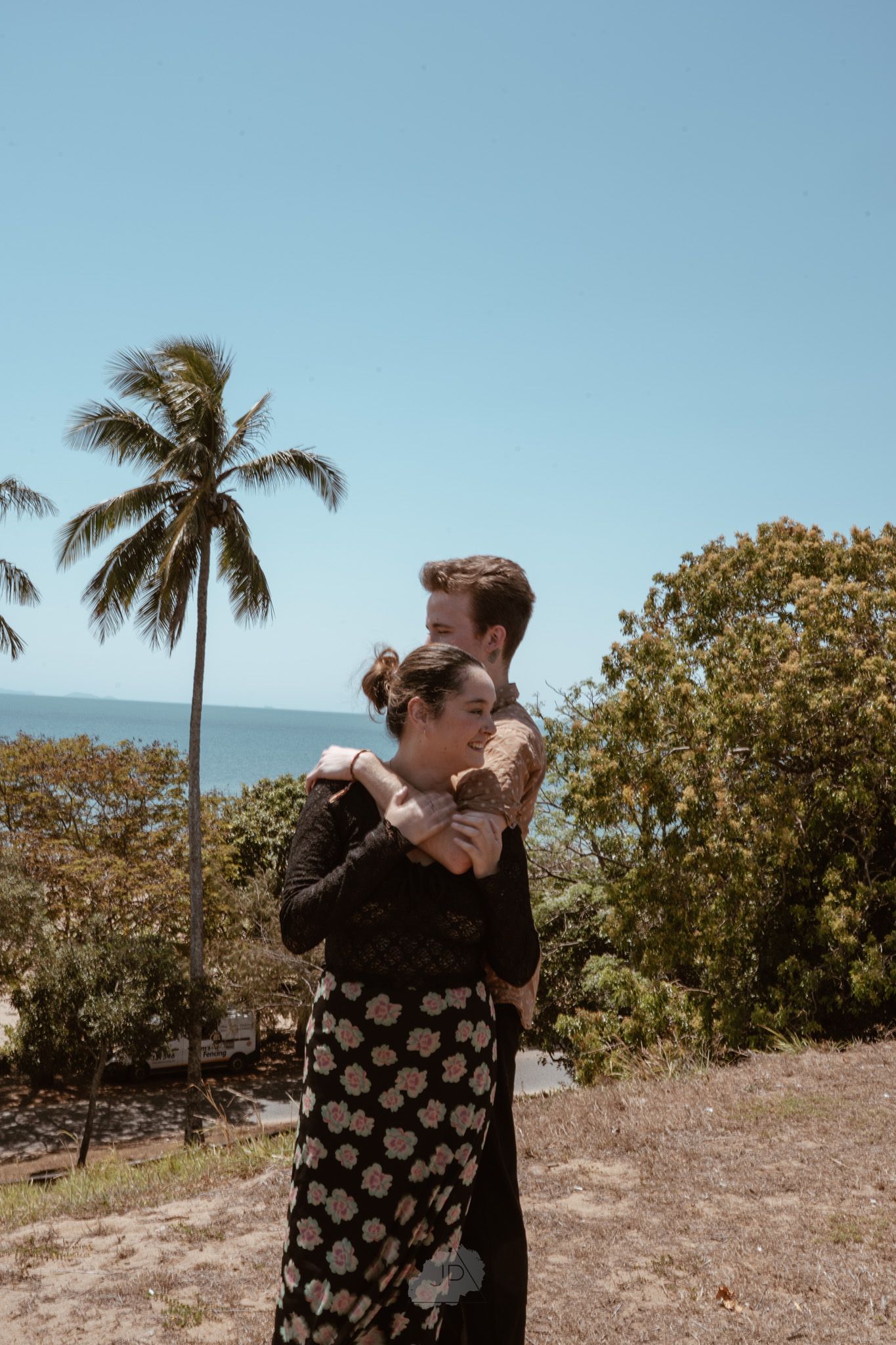 A person in a heart-patterned sweater stands in a tropical landscape with palm trees and ocean views.