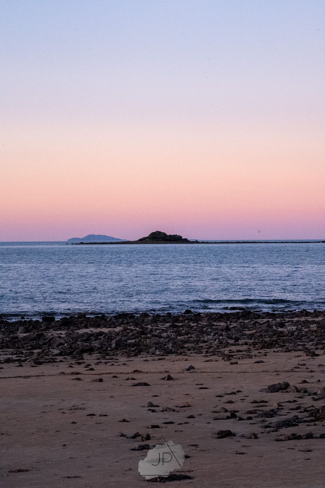 Rocky beach at sunset with pink and purple sky over calm ocean waters and distant mountains on the horizon.