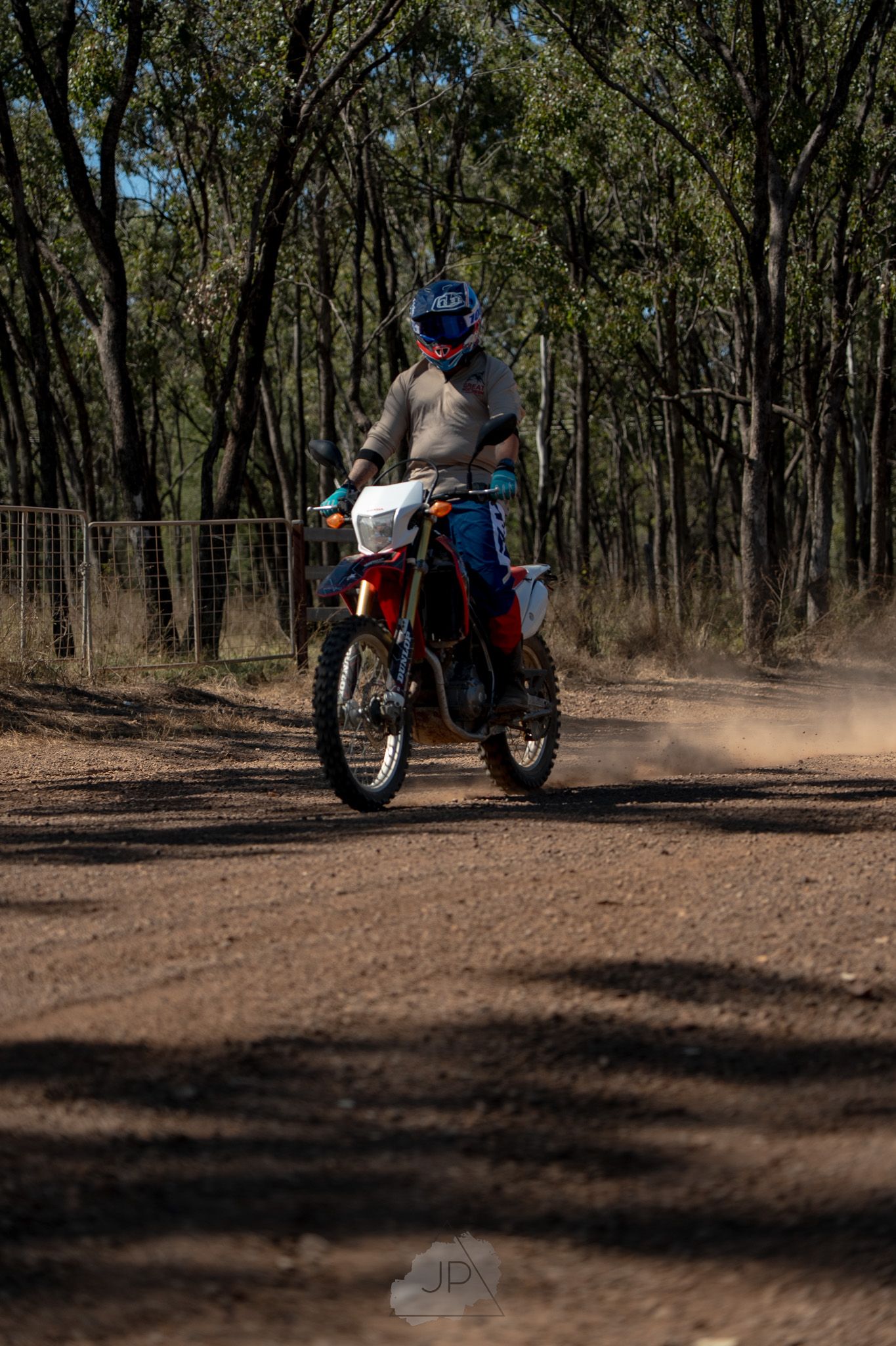 A dirt bike rider kicks up dust while riding through a wooded trail path on an off-road motorcycle.