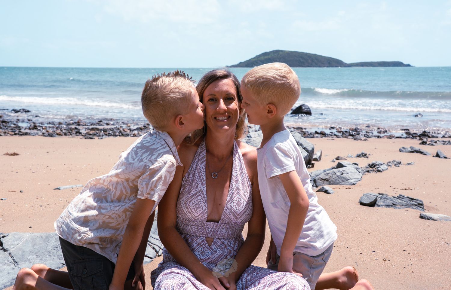 Family sharing a tender moment together on a sunny beach with ocean and rocky coastline in the background.