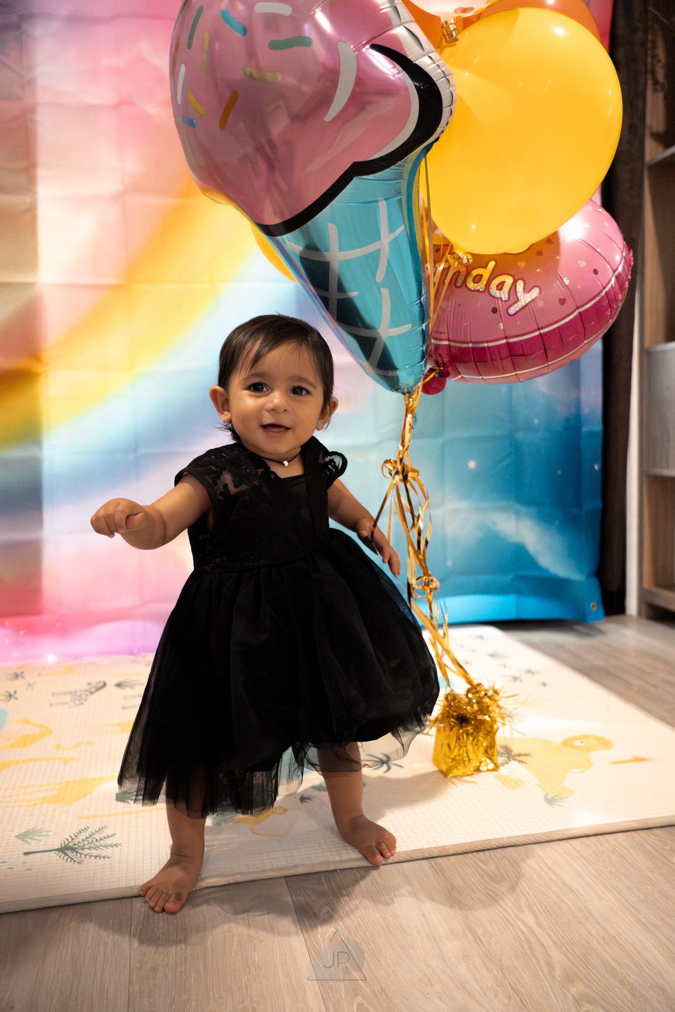 Small child in black tutu dress plays with colorful birthday balloons against pink and blue background.