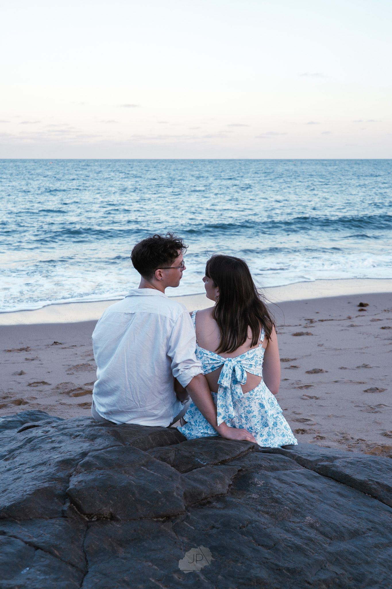 A couple sits together on coastal rocks at sunset overlooking the ocean waves and sandy beach.