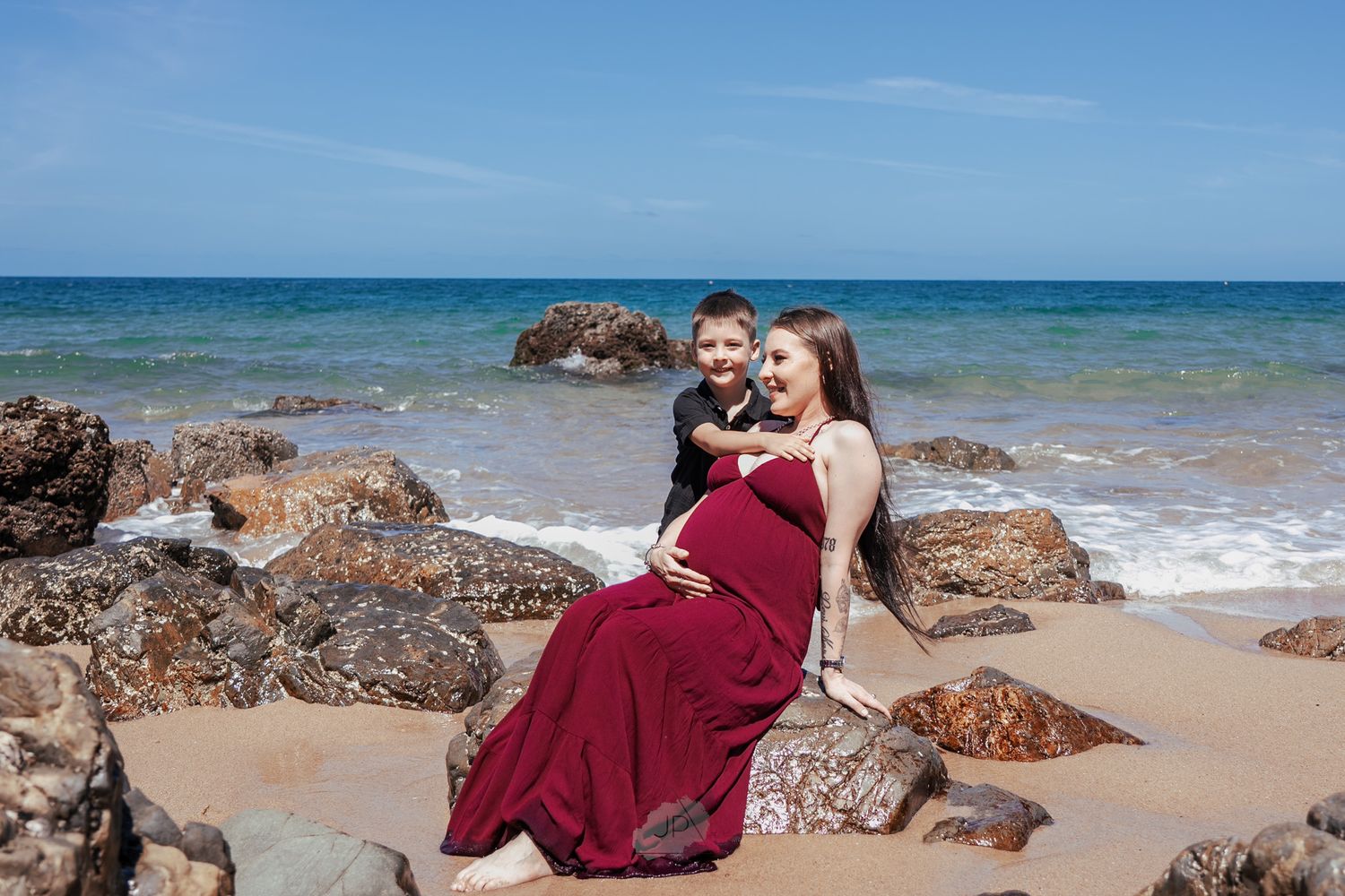 A romantic beach photoshoot features a couple in burgundy attire posing on rocky coastal terrain by the ocean.