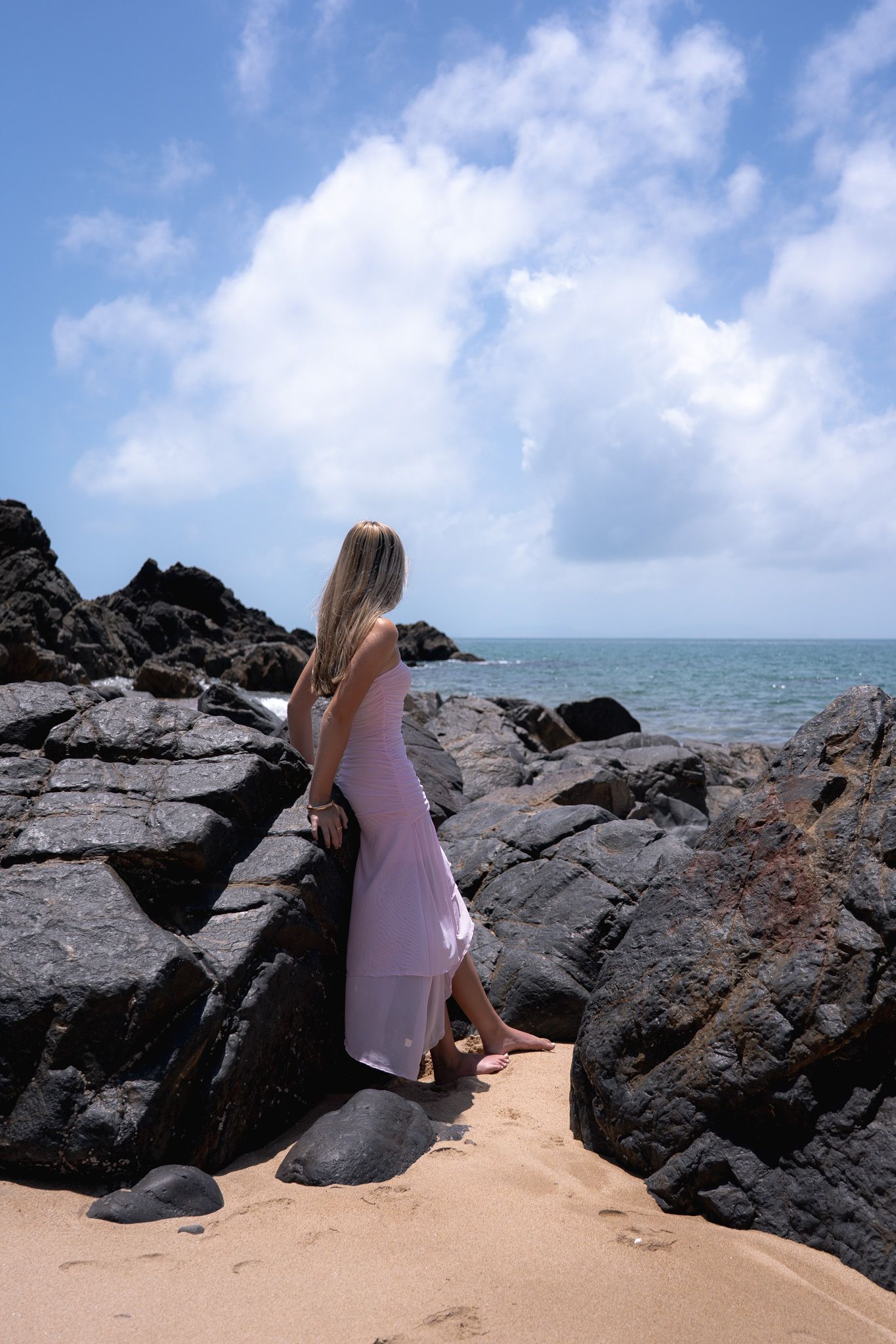 Woman in pink dress stands on sandy beach among dark rocky outcrops against blue ocean and cloudy sky.
