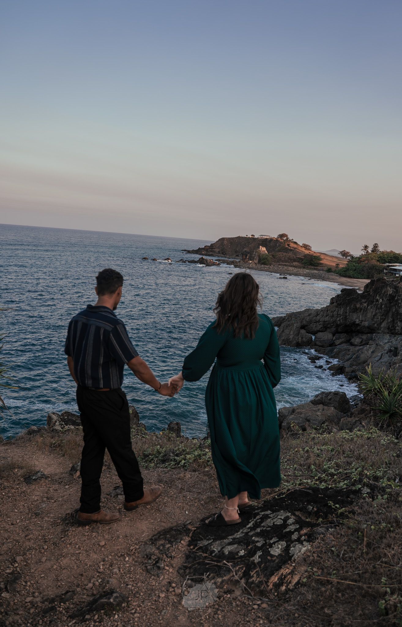 A couple holding hands while standing on rocky coastal cliffs at sunset overlooking the ocean and distant shoreline.