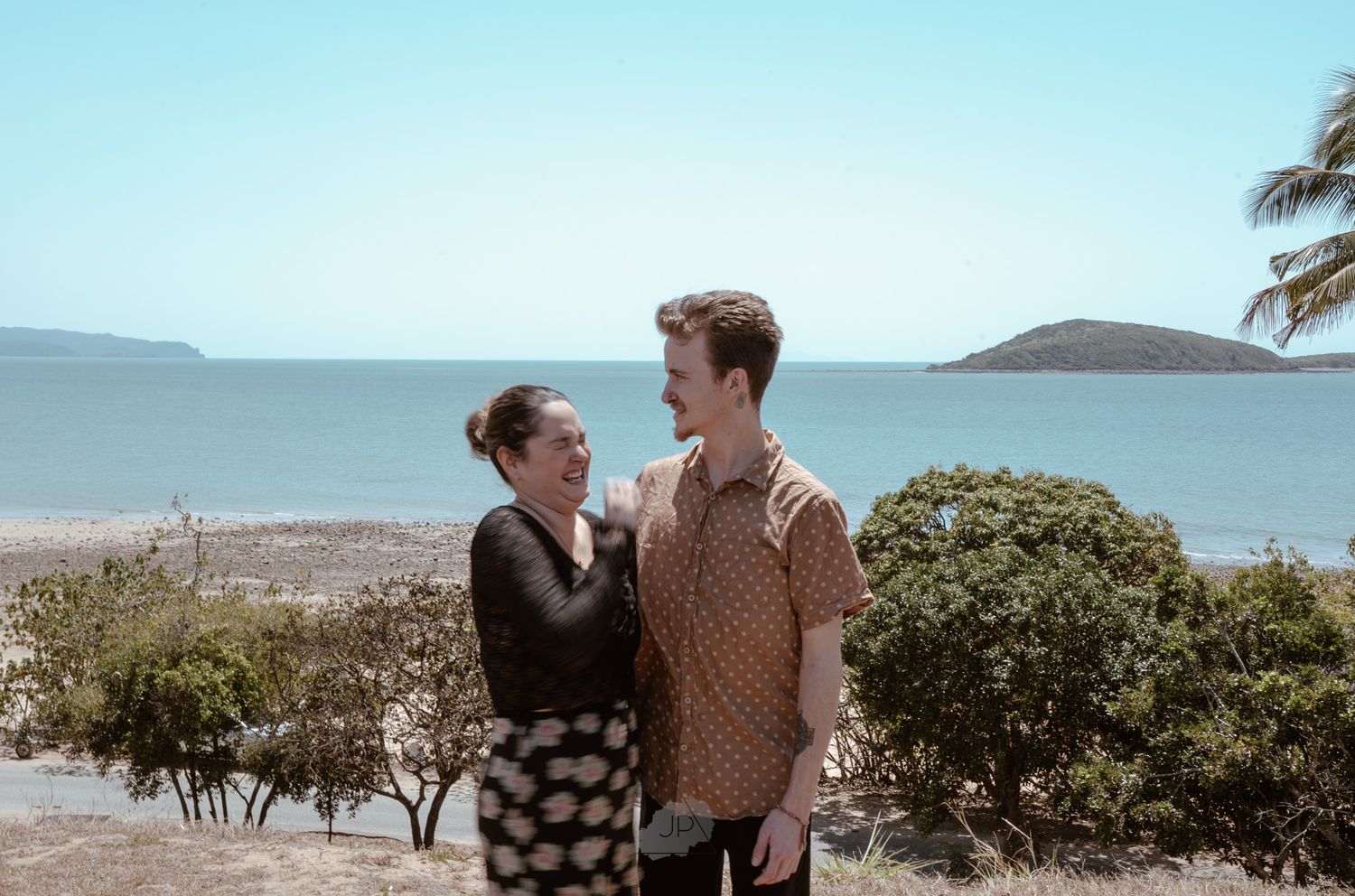 A couple stands together on a scenic beach overlook with palm trees and ocean views in the background.