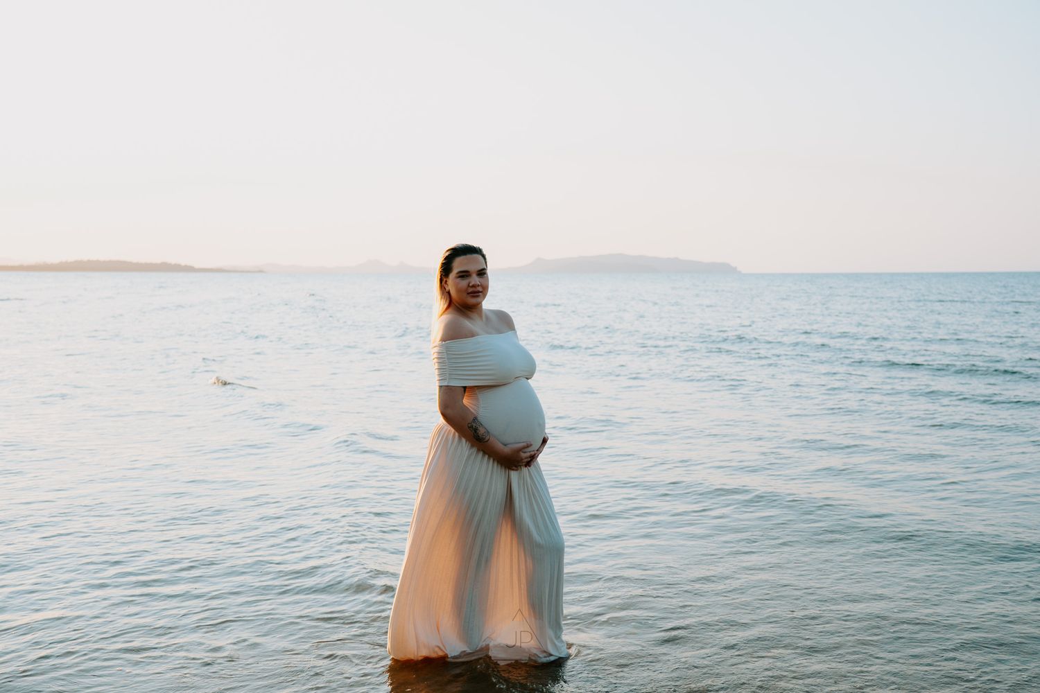 A pregnant person in a flowing white dress stands at the ocean's edge during sunset, cradling their belly.