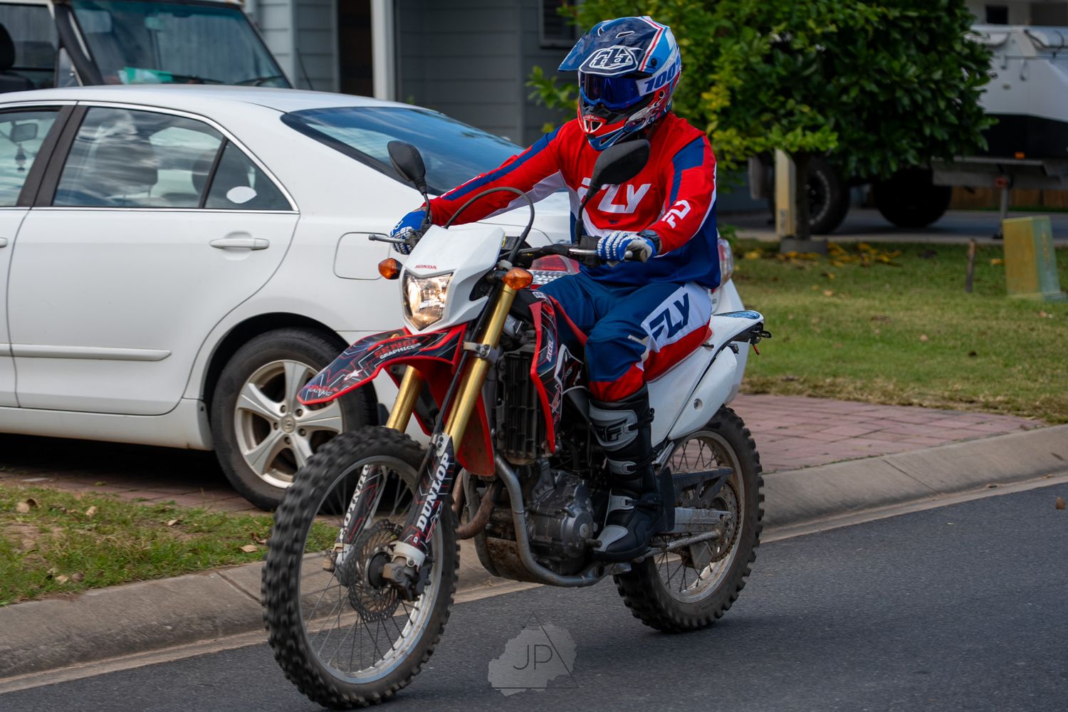 A Honda CR500 dirt bike with red and blue color scheme parked in front of trees and houses on a residential street.