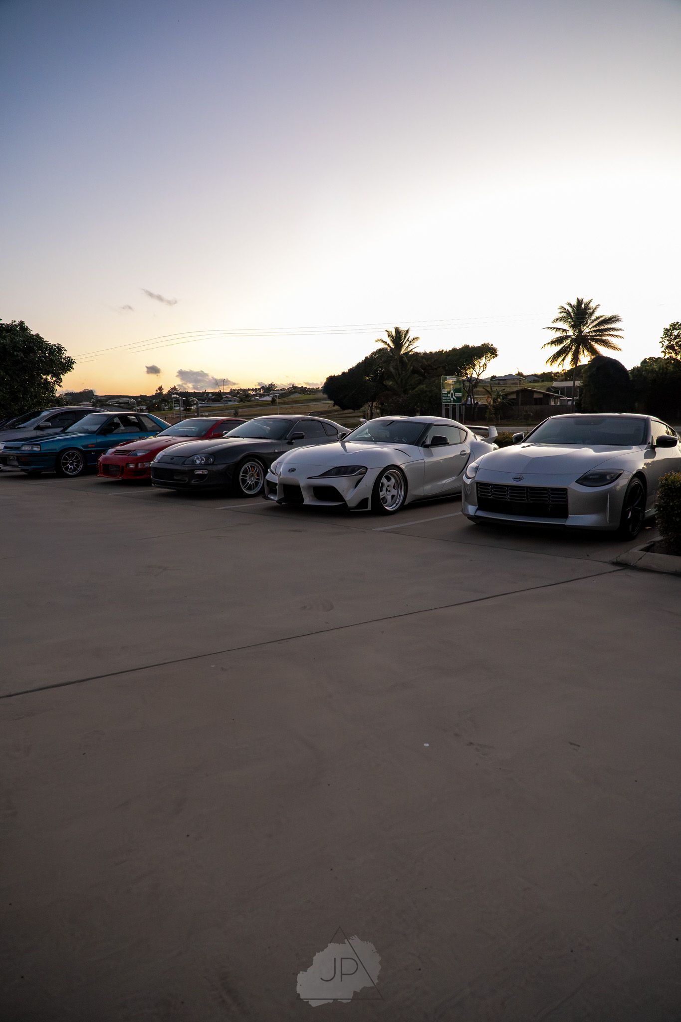 Row of luxury sports cars including Ferraris and Porsches parked at sunset in a parking lot.