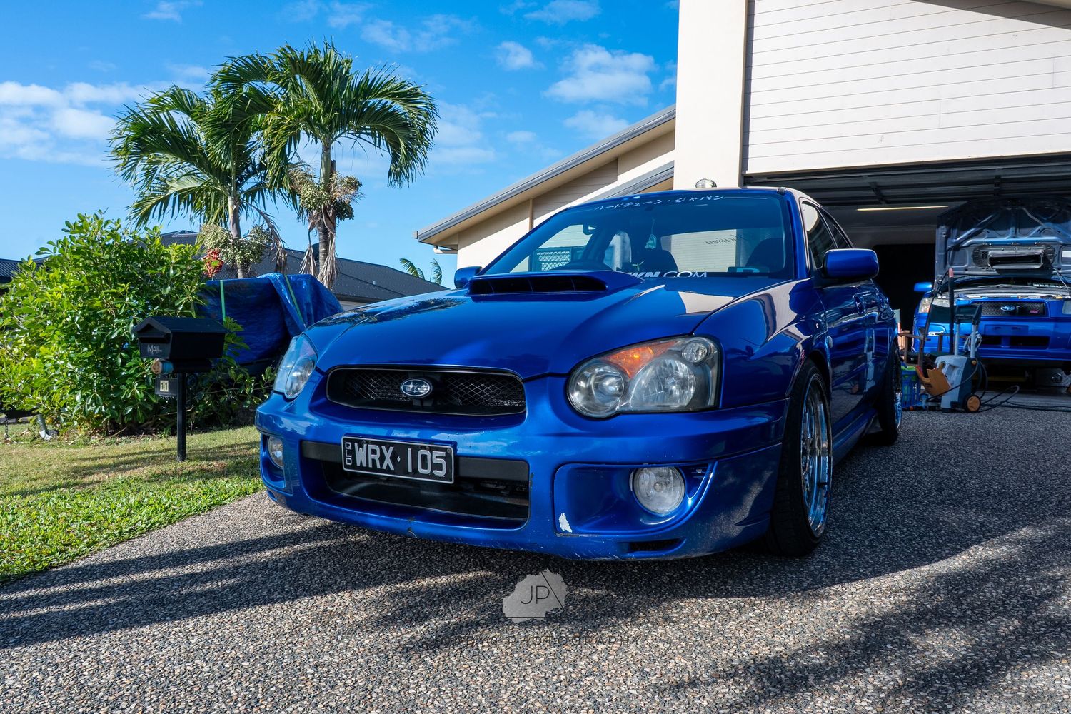 A bright blue Subaru WRX STI is shown from multiple angles parked in a gravel driveway against a sunny sky.