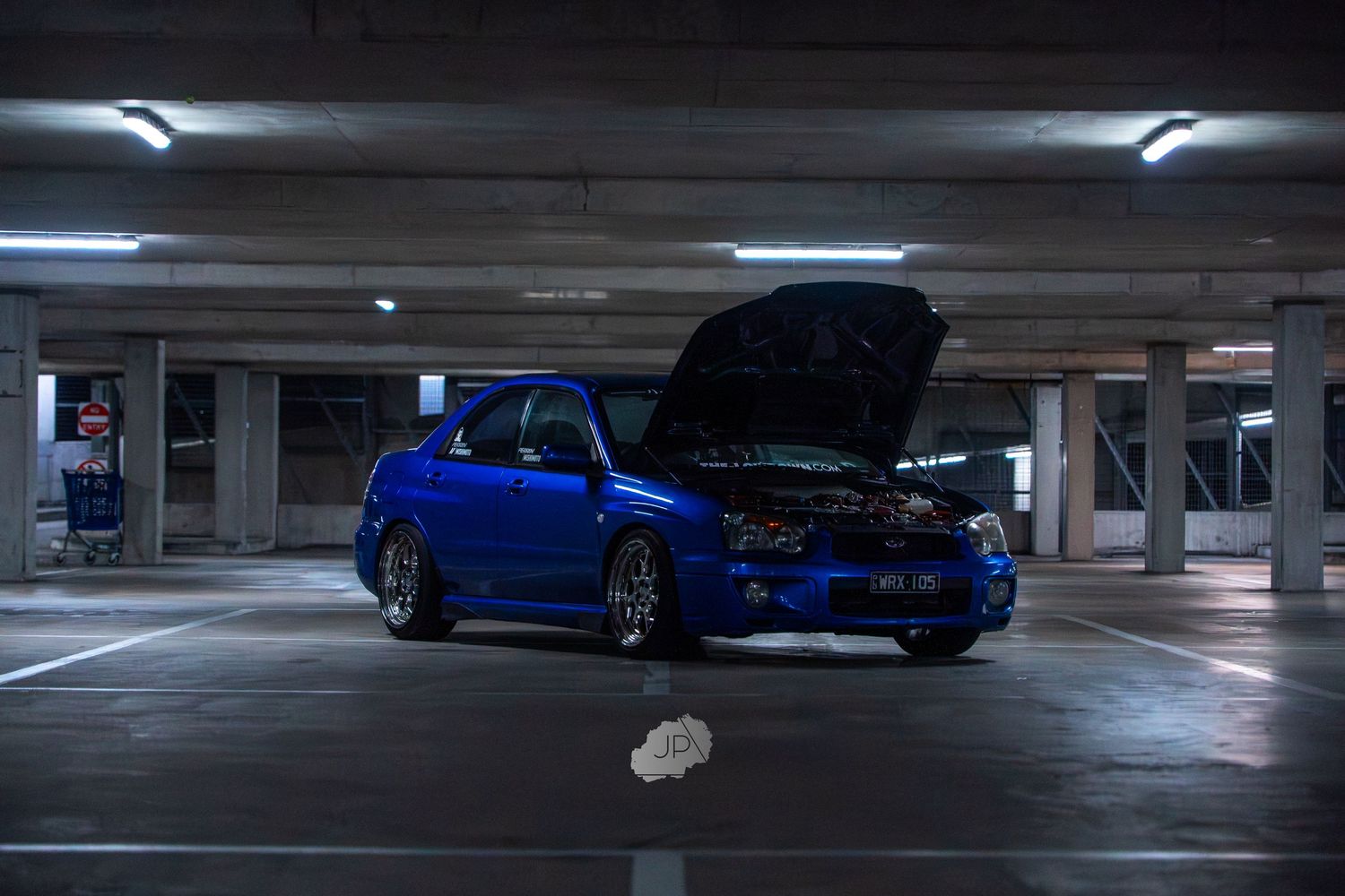 Blue Subaru WRX STI with open hood parked in dimly lit parking garage showing performance upgrades.