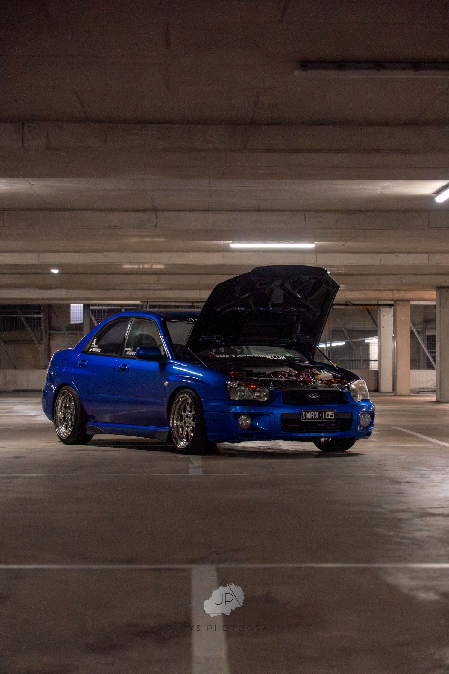 A bright blue modified car with an open hood sits parked in a dimly lit concrete parking garage.