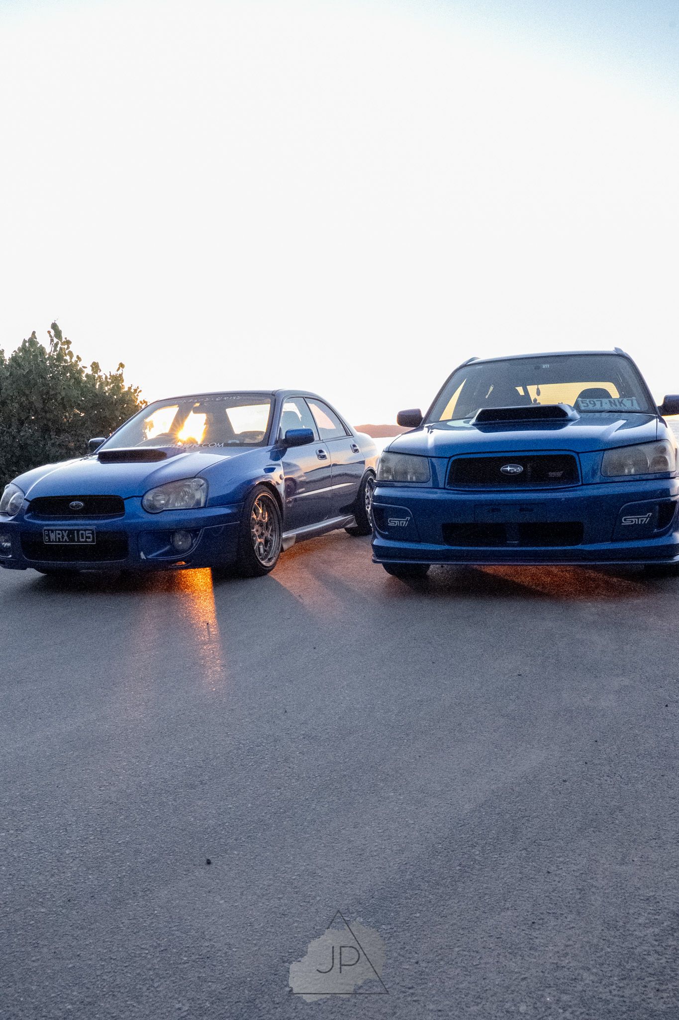 Two blue Subaru WRX performance cars parked side by side at sunset with dramatic lighting on the ground.