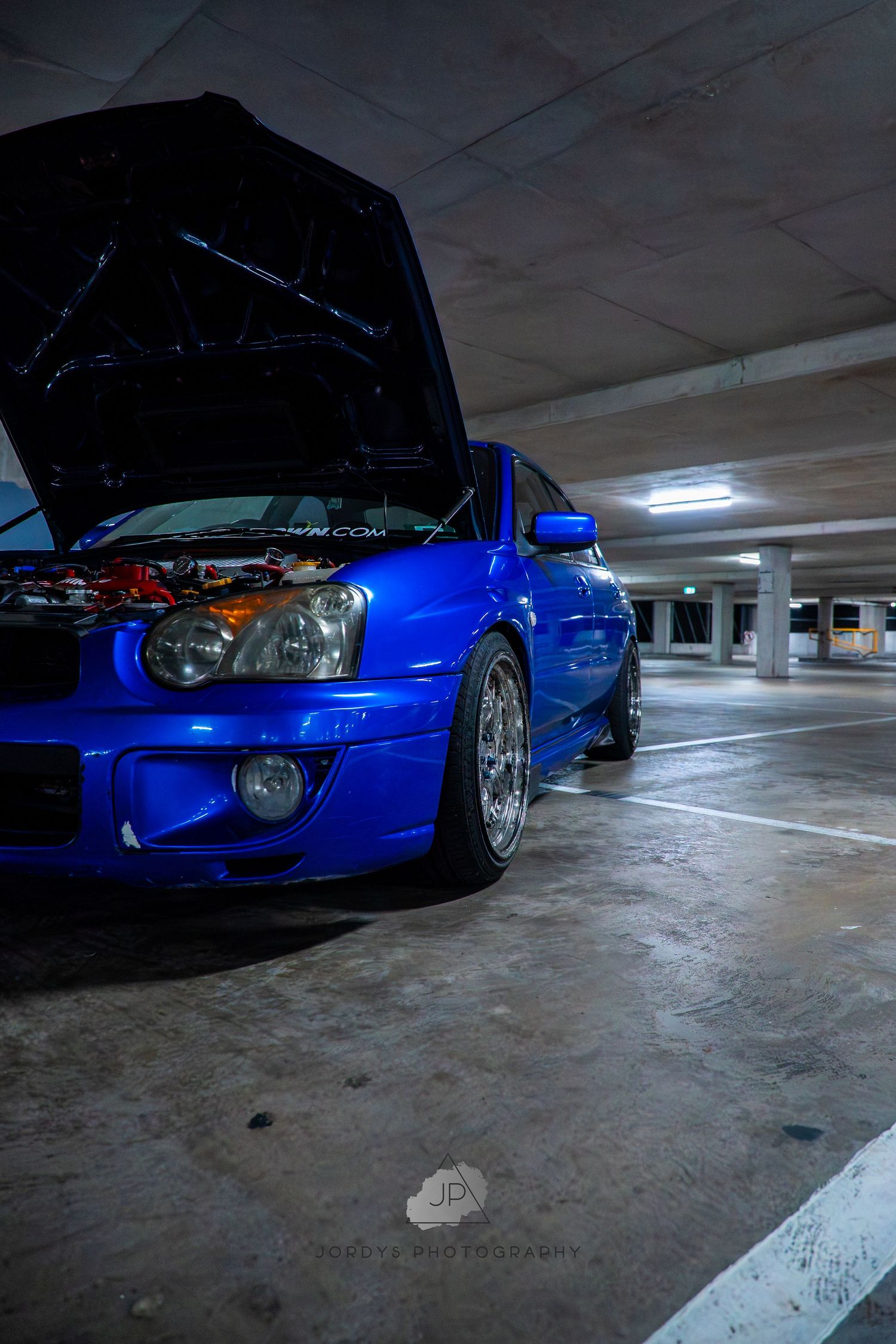 A bright blue lowered sports car with open hood sits in a dimly lit parking garage from multiple angles.