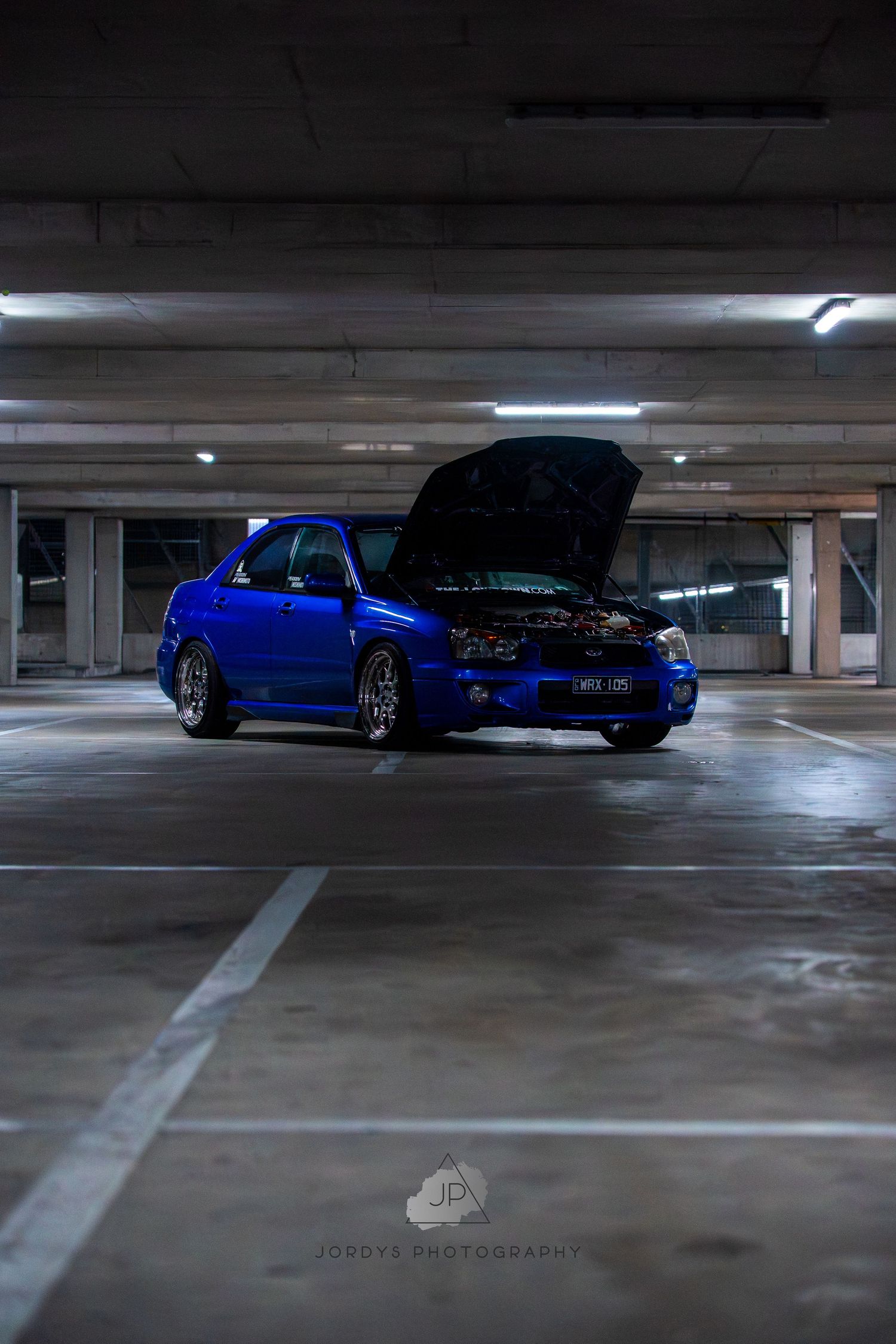 A stylish blue sports car is parked in a dimly lit concrete parking garage.