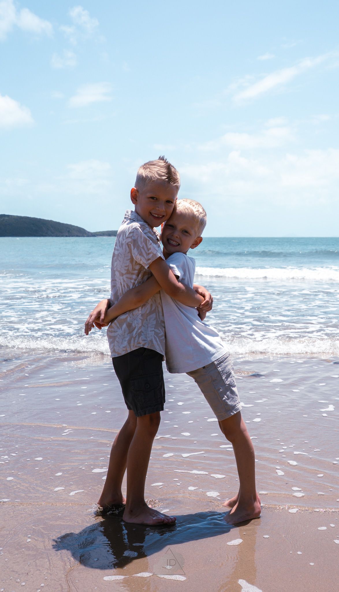 Two children playing and hugging together on a sunny tropical beach with waves and mountains in the background.