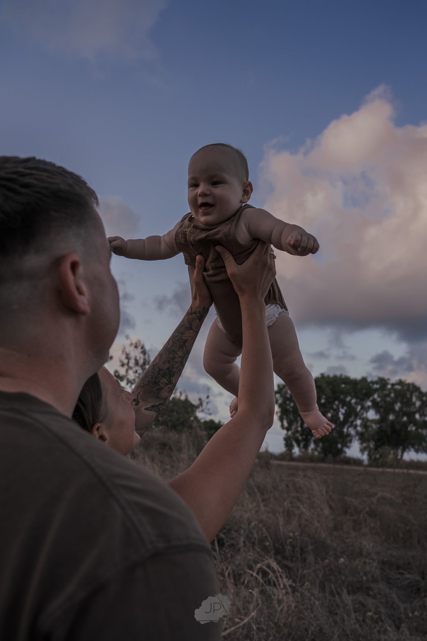 A person lifts and plays with a baby against a cloudy sky backdrop during sunset.