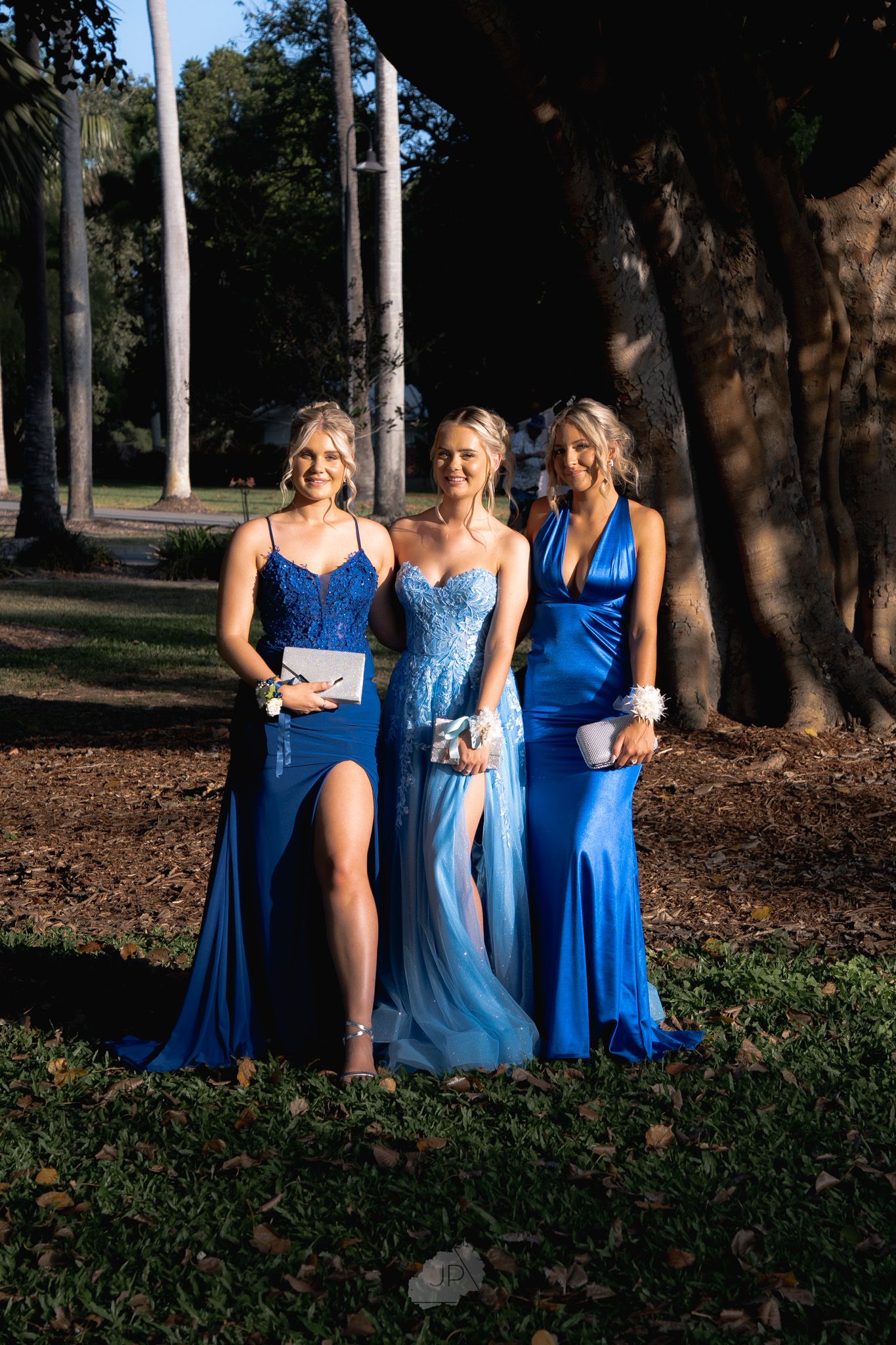 A group in coordinating blue dresses poses together in a wooded outdoor setting during a formal event.