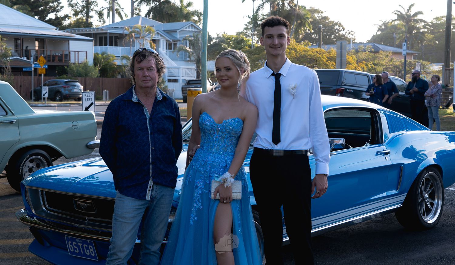 A group of people pose together at a classic car show featuring vintage blue automobiles in an outdoor setting.