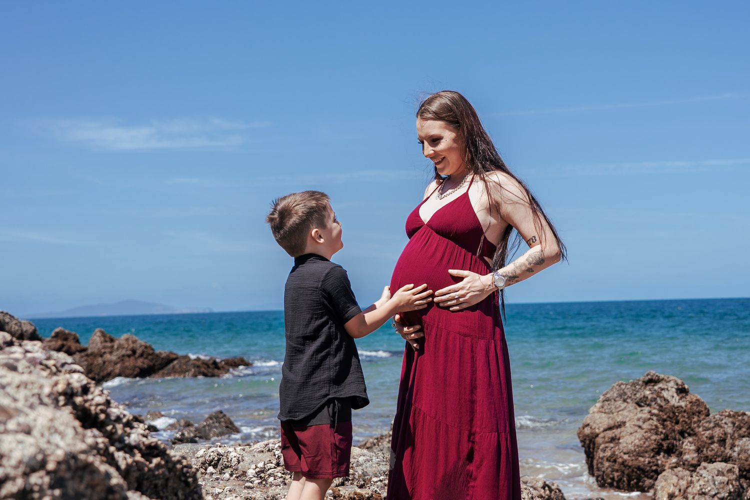 A couple poses for engagement photos on a rocky beach with turquoise ocean waters and blue skies in the background.