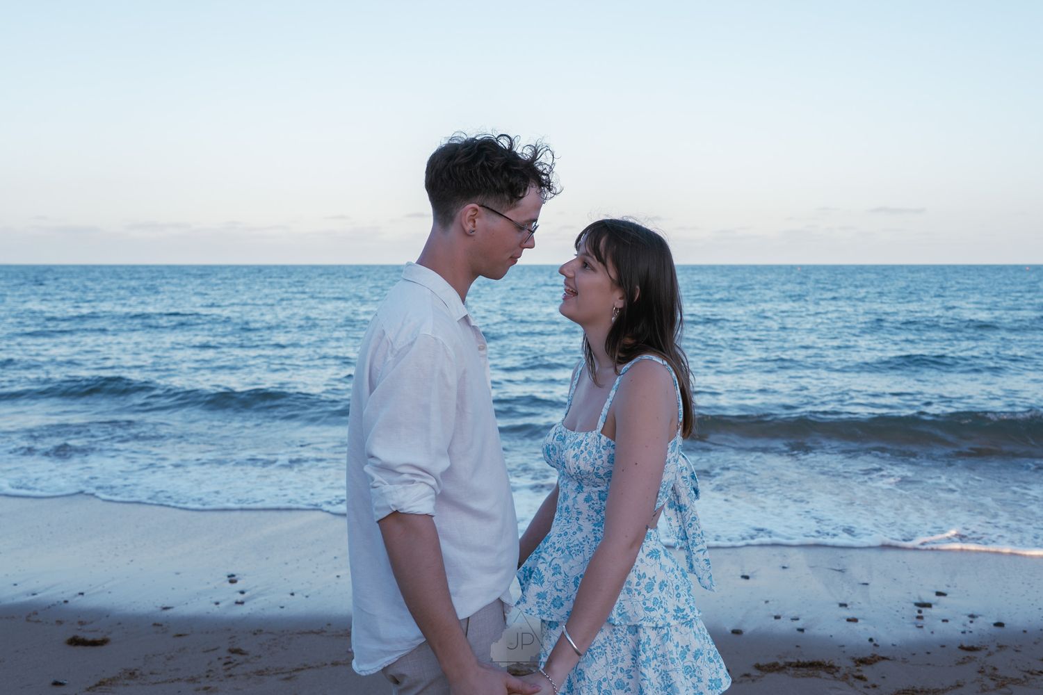 A romantic couple embraces on a scenic beach at sunset with ocean waves in the background.
