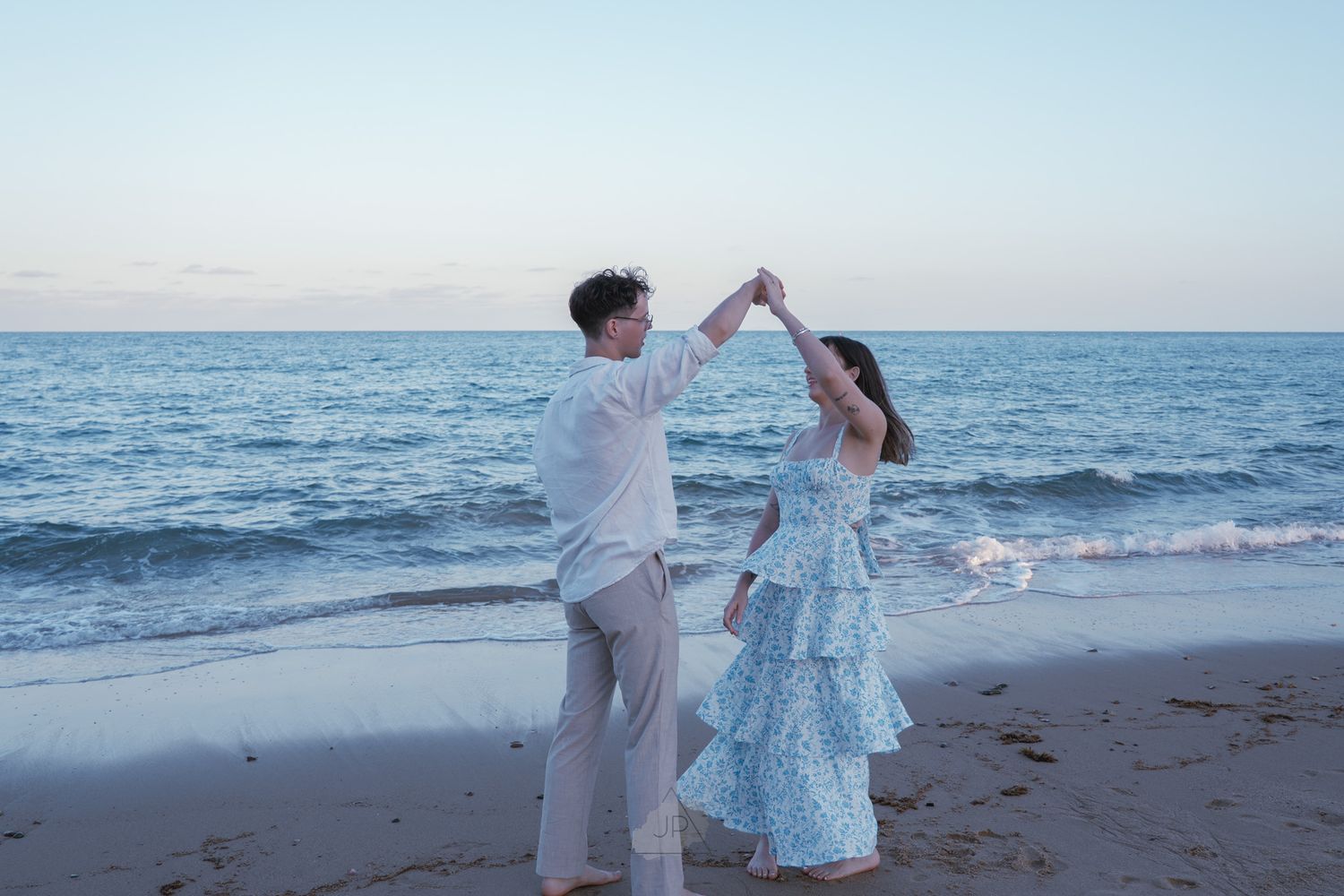 A couple dances together on a scenic beach at sunset with ocean waves in the background.