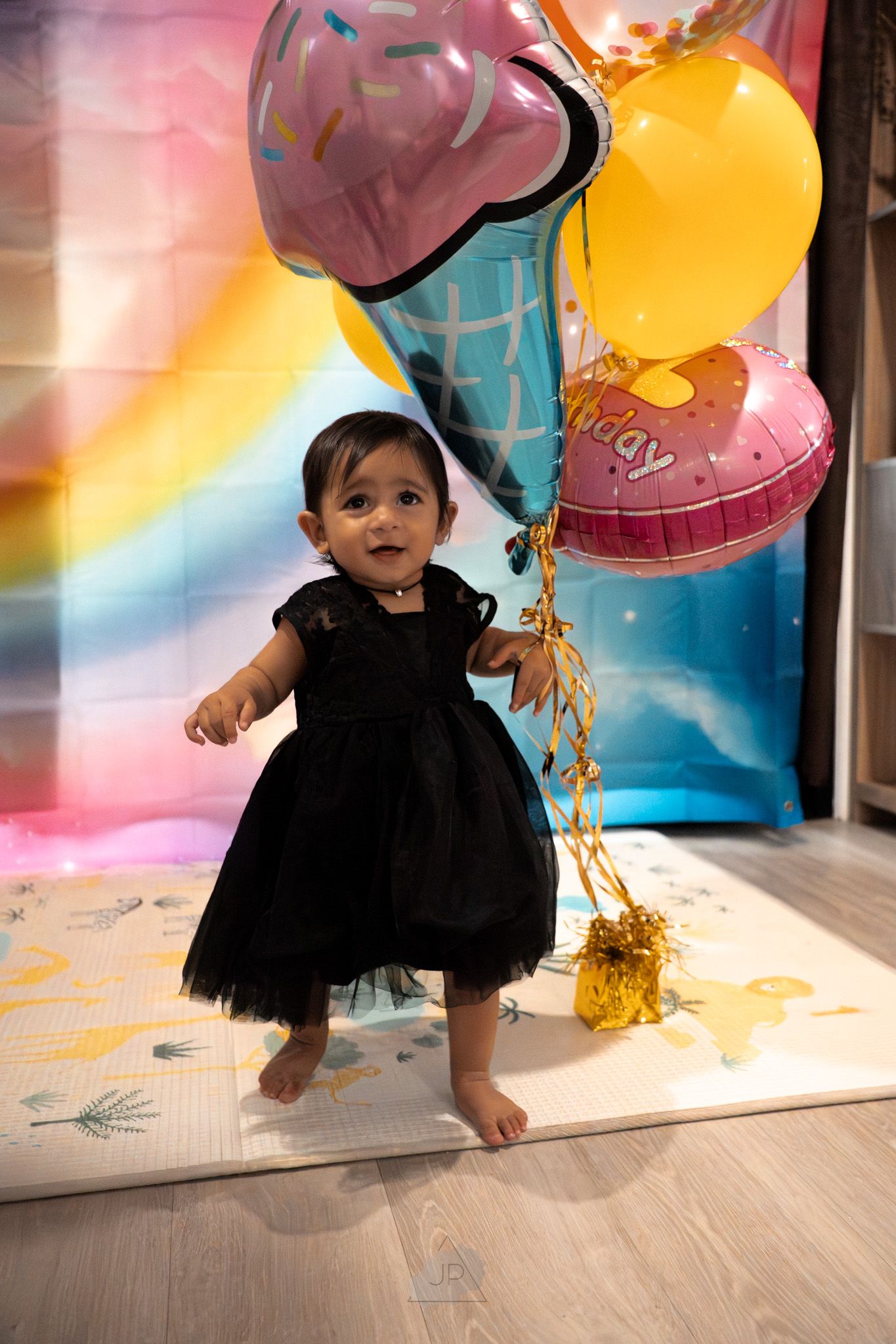 A small child in a black tutu dress plays with colorful balloons against a dreamy pastel background.