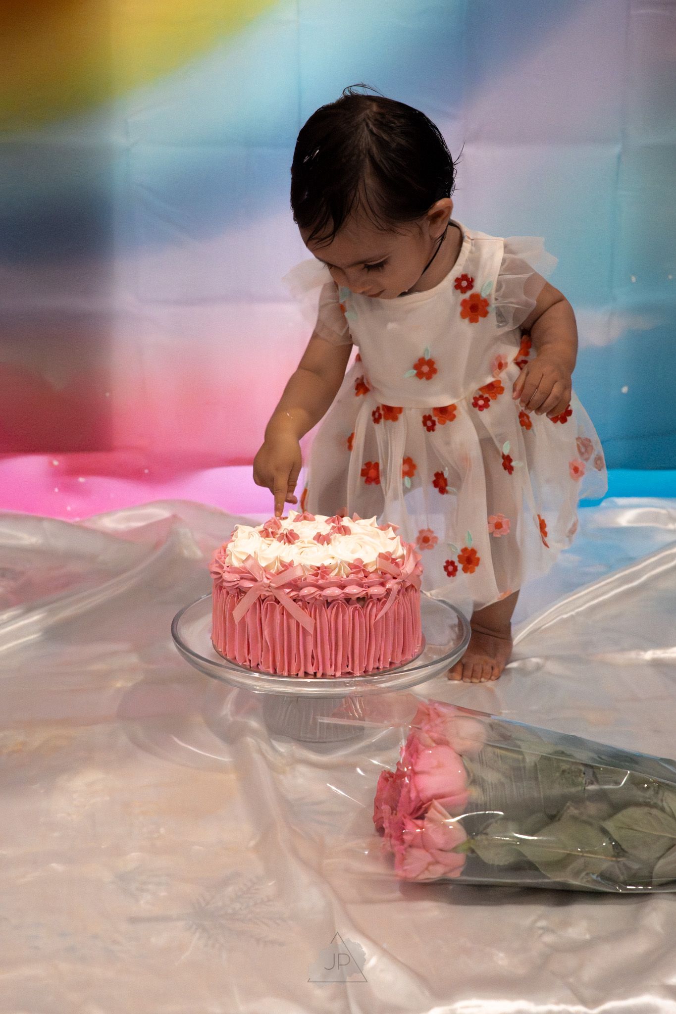 A baby in a white dress with red flowers explores a pink birthday cake during a cake smash photo session.