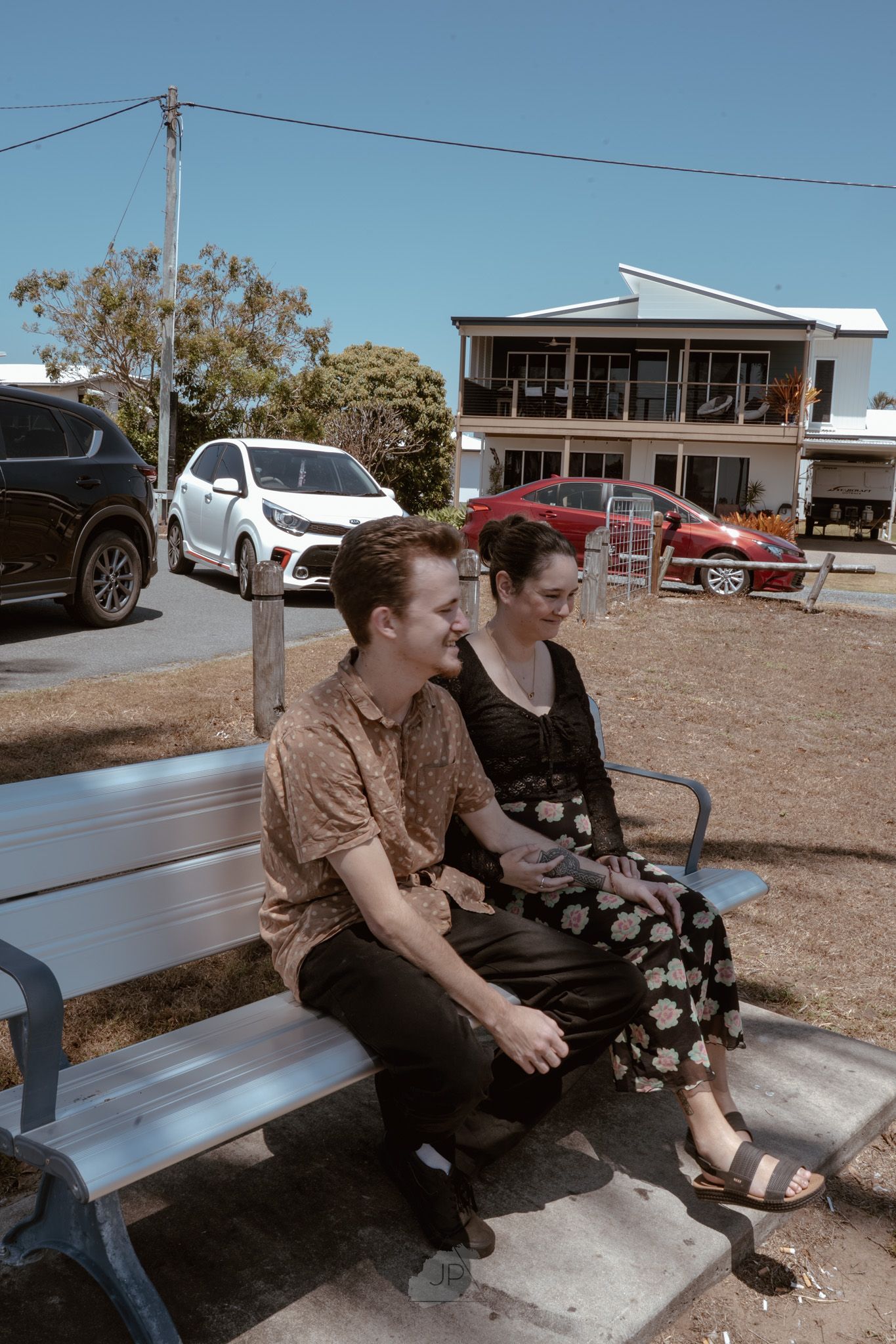 Two people share a conversation while sitting on a metal bench outdoors on a sunny day.