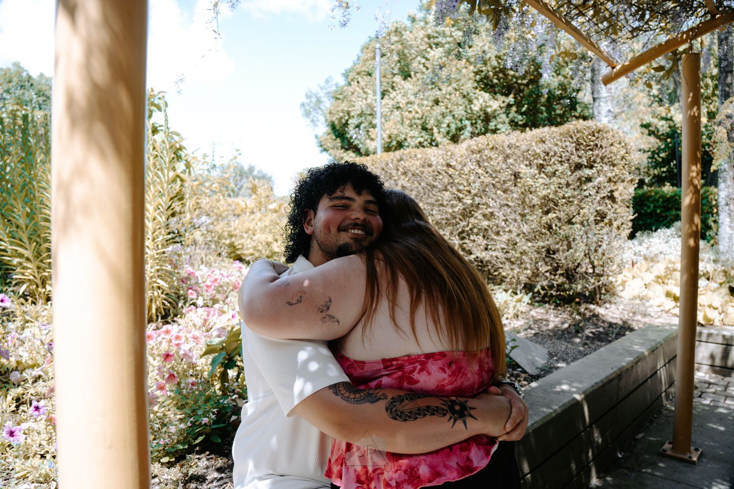 Two people share a tender embrace under a wooden pergola in a sunlit garden setting.