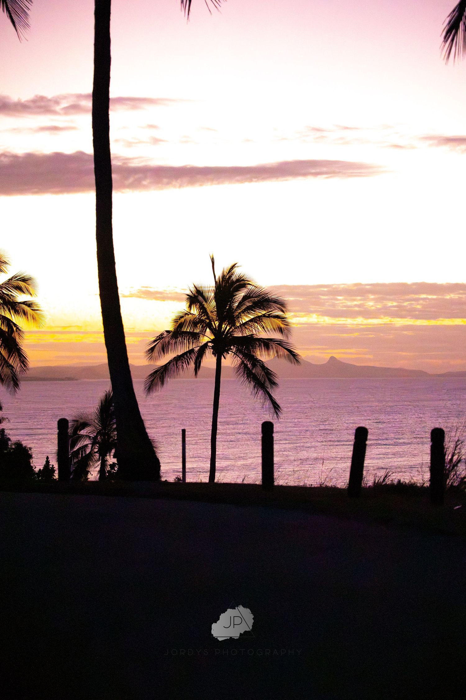 Silhouettes of palm trees against vibrant orange and pink sunset sky over ocean horizon with distant islands.