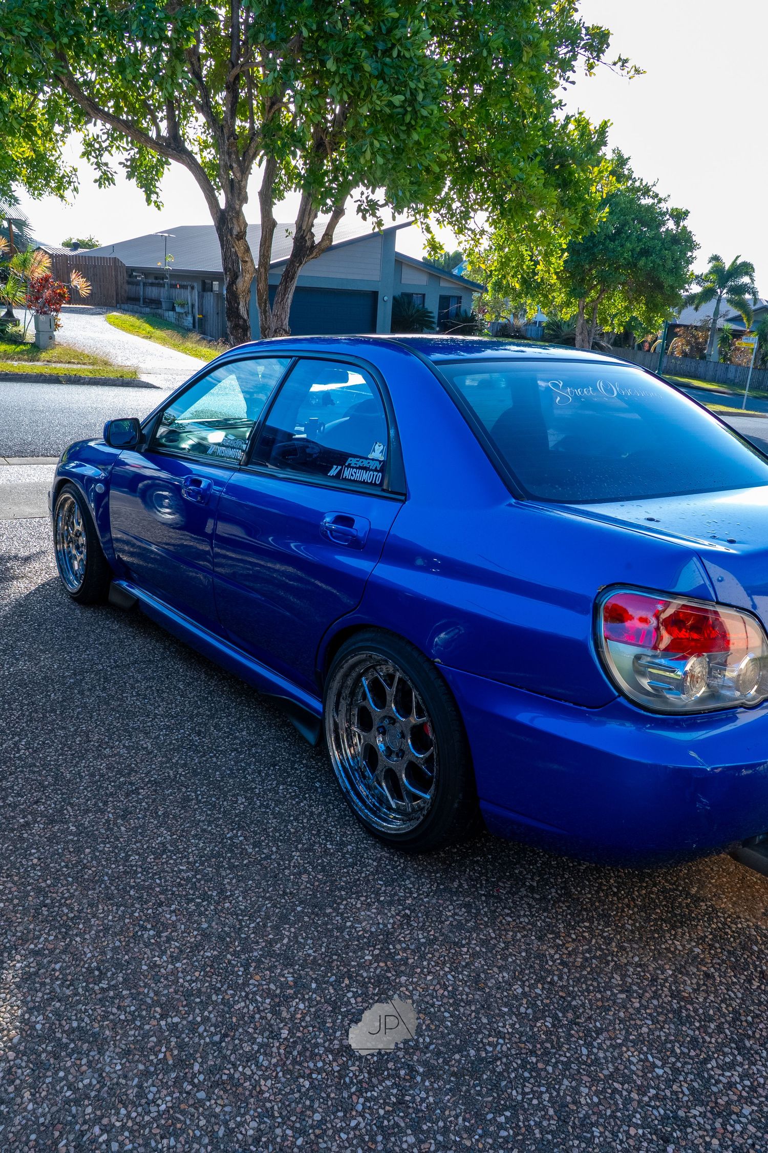 Low profile Japanese sports car in metallic blue shown at multiple angles on gravel surface under trees.