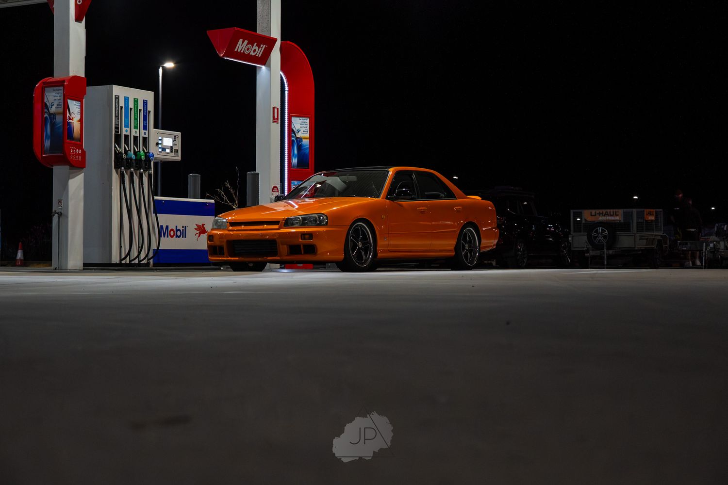 Orange sports car parked at a gas station at night with bright lighting and reflective surfaces.