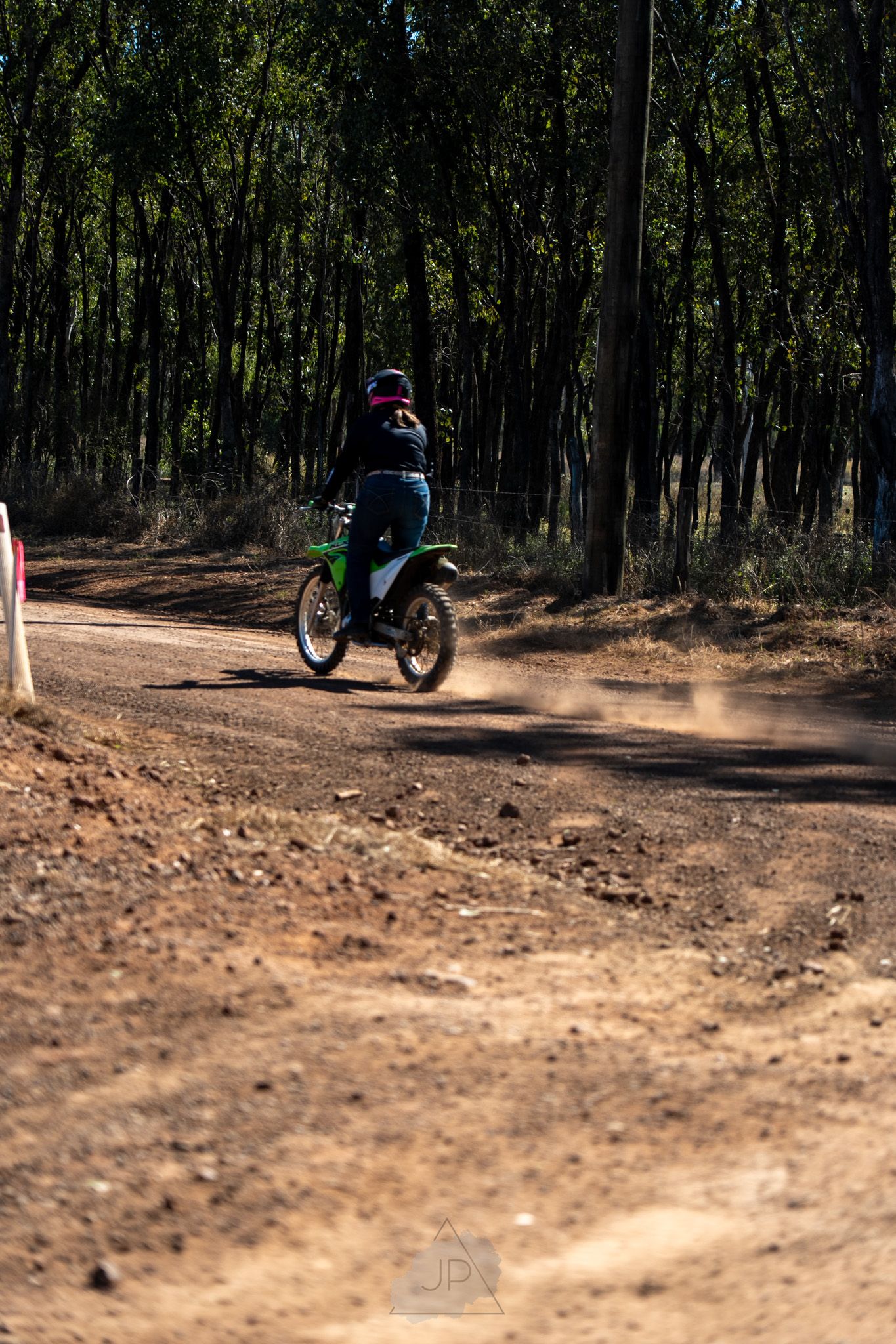 Mountain biker navigates a curvy dirt trail through a wooded forest area, creating dust clouds behind the tires.