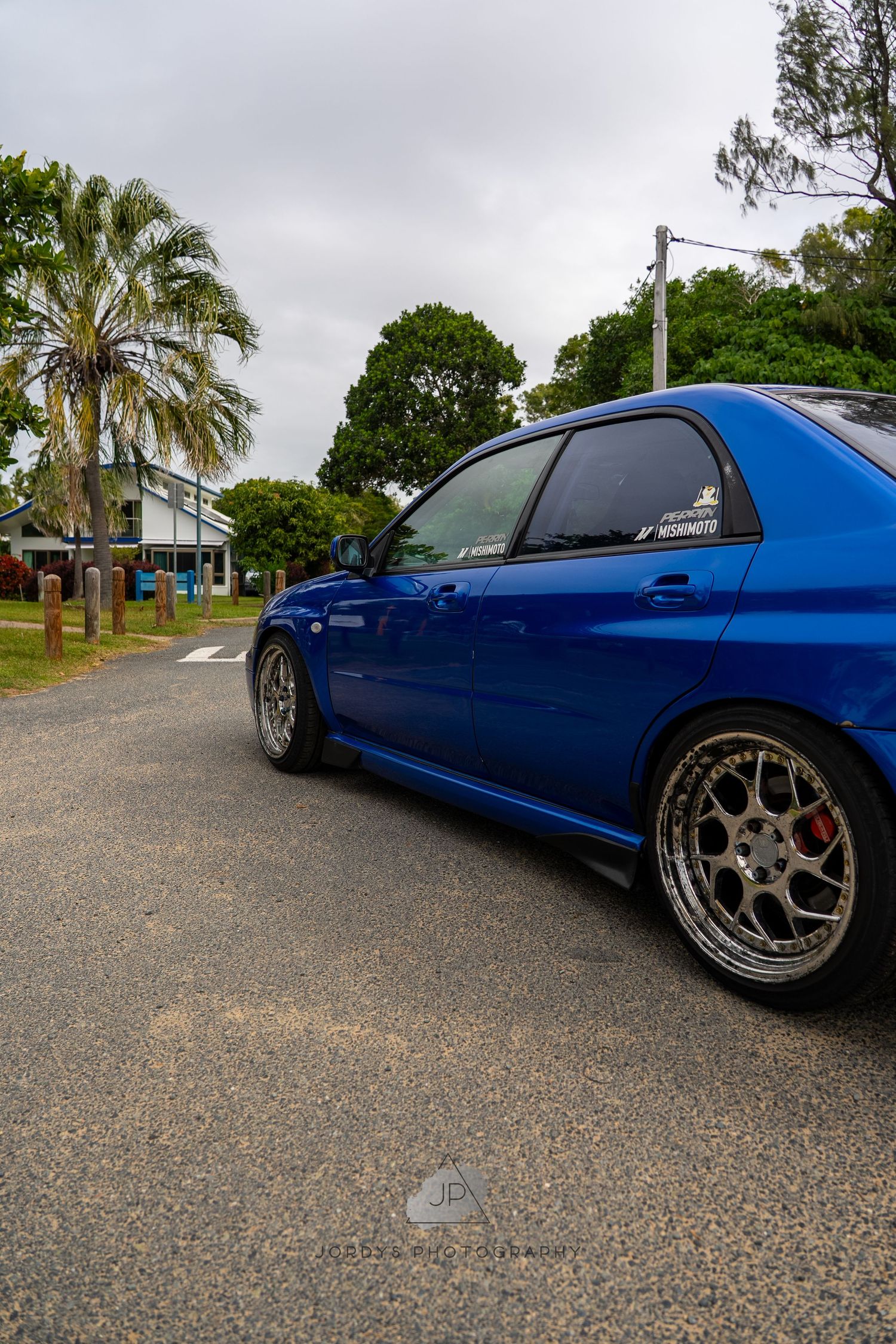 A lowered bright blue sports car with custom wheels parked on a gravel surface with palm trees in the background.