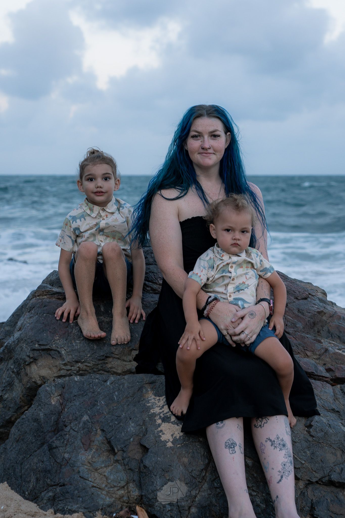 Family sitting together on coastal rocks at dusk with stormy ocean waves and cloudy sky in background.