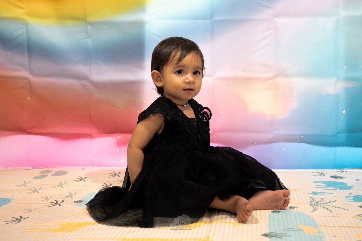 A small child in a black dress sits on a colorful backdrop with soft pastel lighting effects.