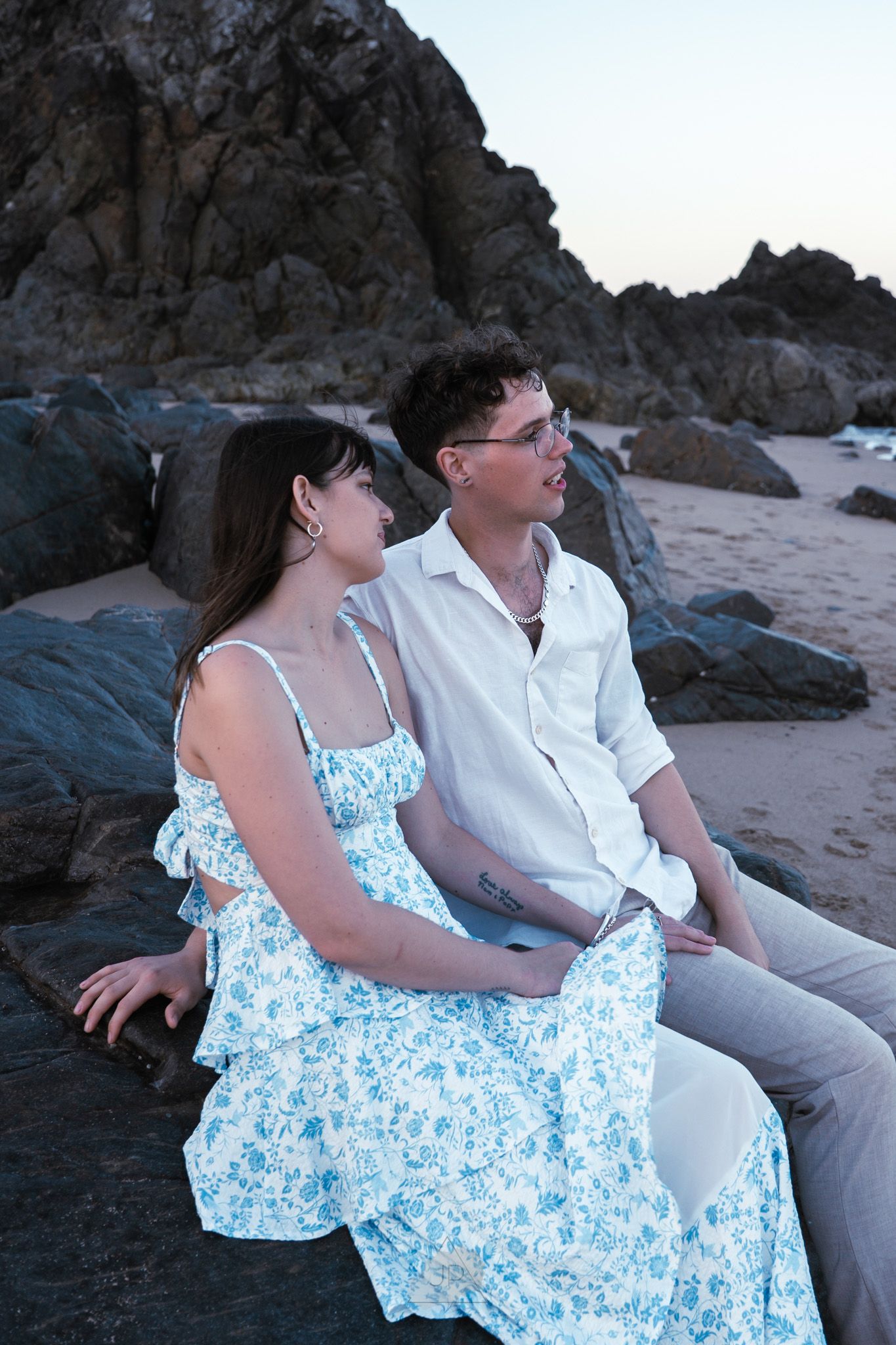 A couple sits together on a rocky beach at sunset, with the woman wearing a blue and white floral dress.