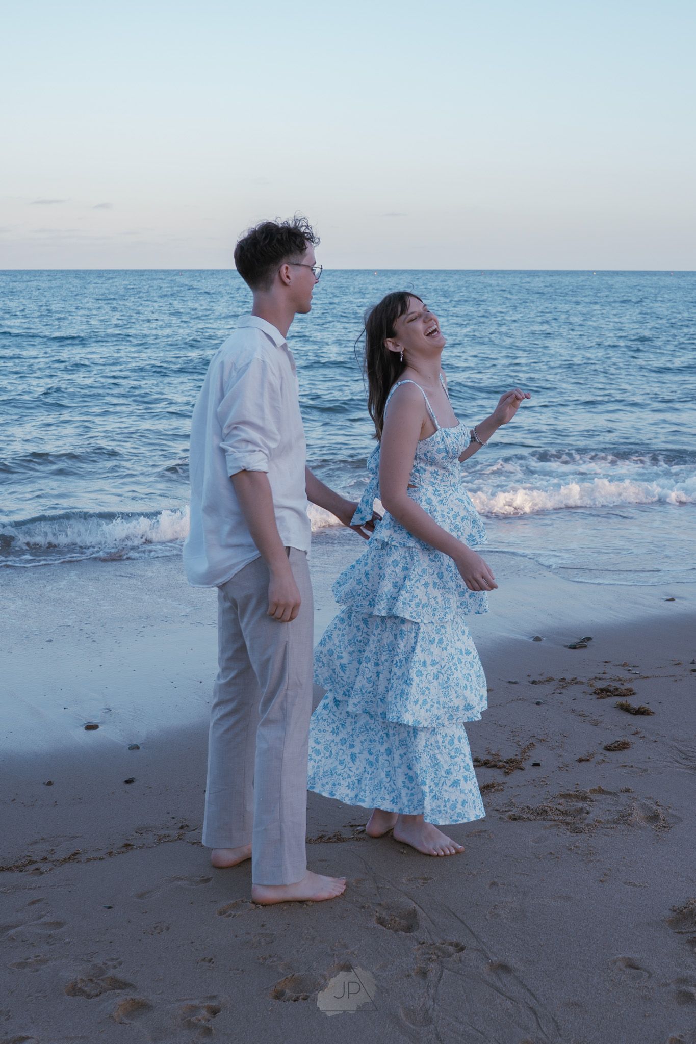 Two young children in light colored outfits play together at the edge of the ocean during sunset.