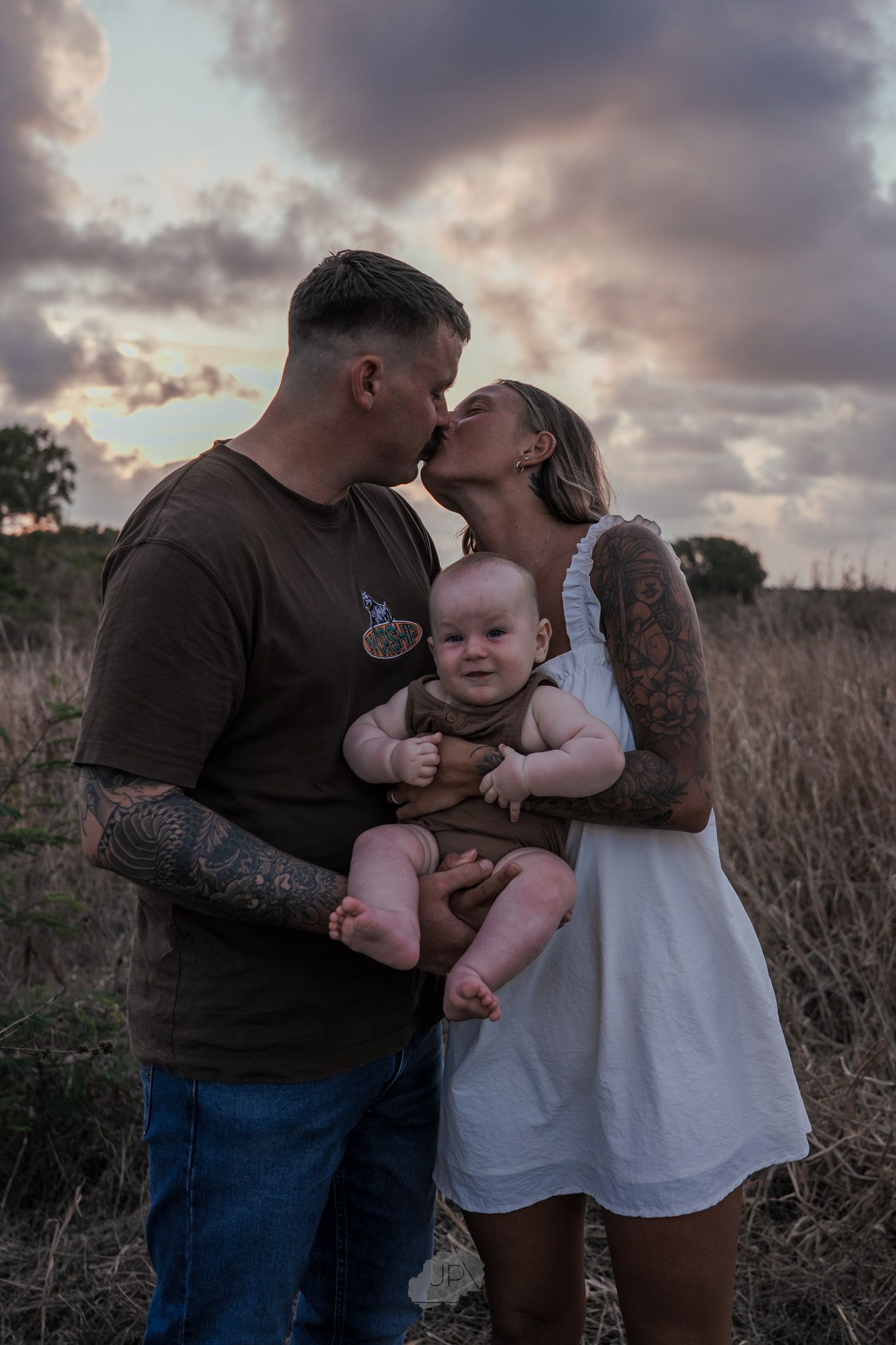 A couple shares a tender kiss while holding their baby in a scenic outdoor field at sunset.