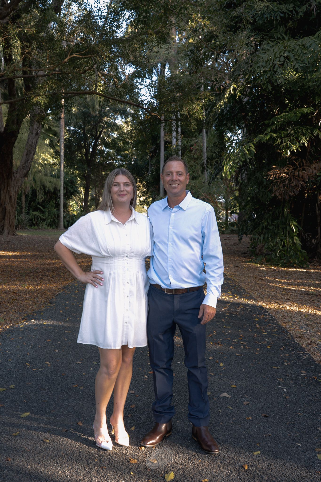 A couple poses together outdoors on a path surrounded by trees and fallen autumn leaves in a park setting.